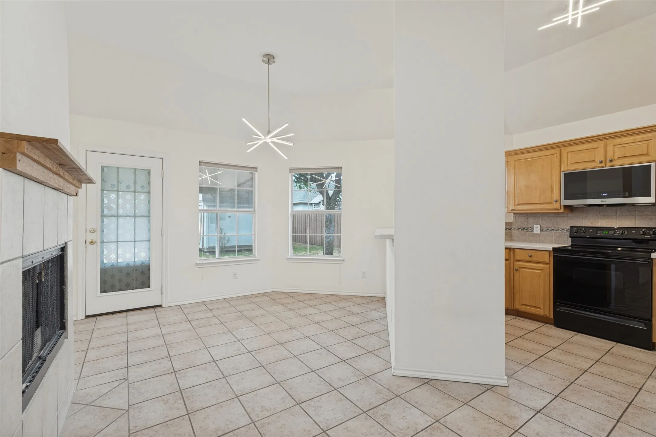 Vaulted ceilings with a contemporary pendant light fixture, tile flooring, a fireplace with a wood mantle, and a kitchen with wood cabinetry and black appliances