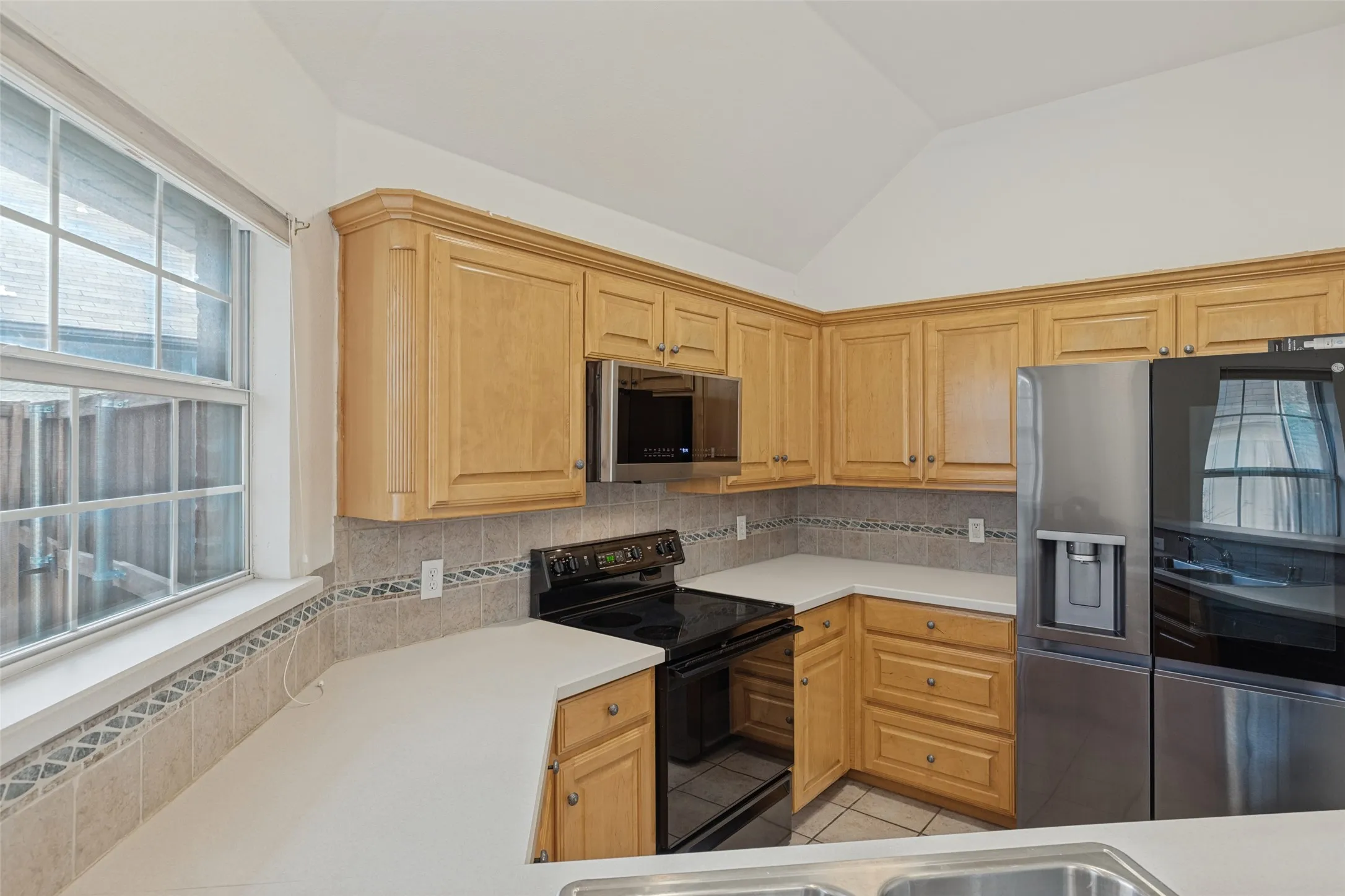 Kitchen featuring light wood cabinetry, white countertops, tile backsplash, stainless steel refrigerator, and integrated cooktop oven