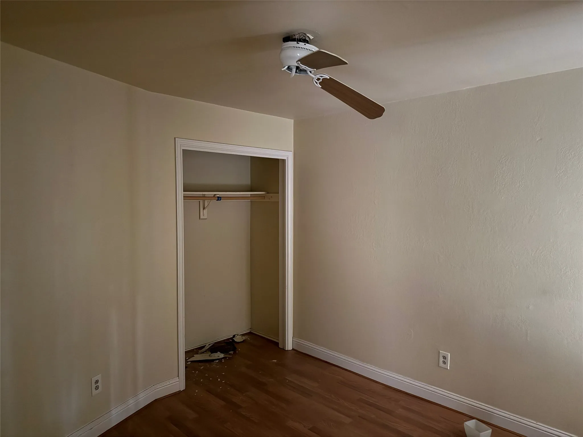 Wood-finish flooring with white trim, ceiling fan with wood-finish blades, and an open closet with a single hanging rod