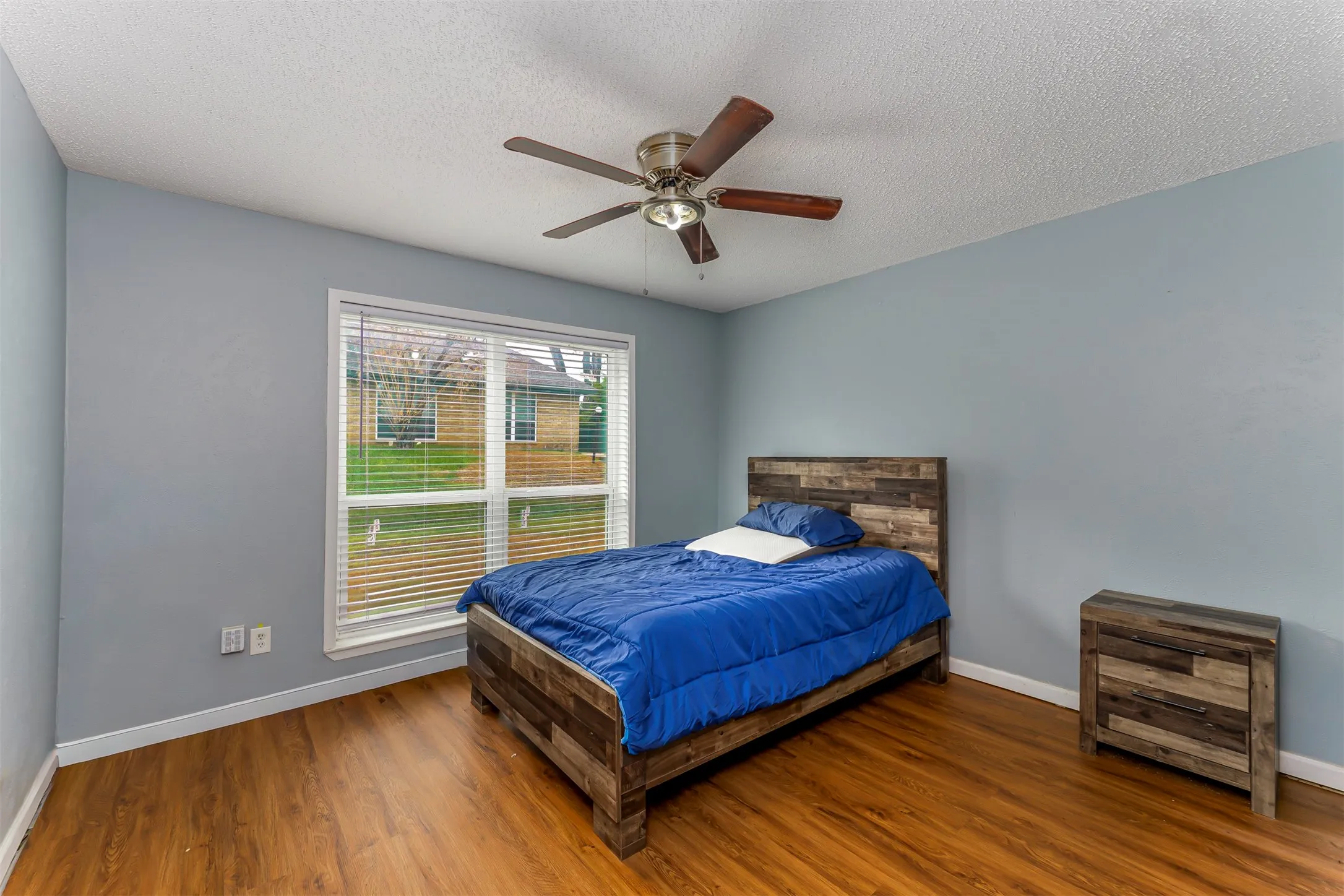 Wood-finish flooring, light blue wall paint, a ceiling fan with light fixture, and a window with horizontal blinds