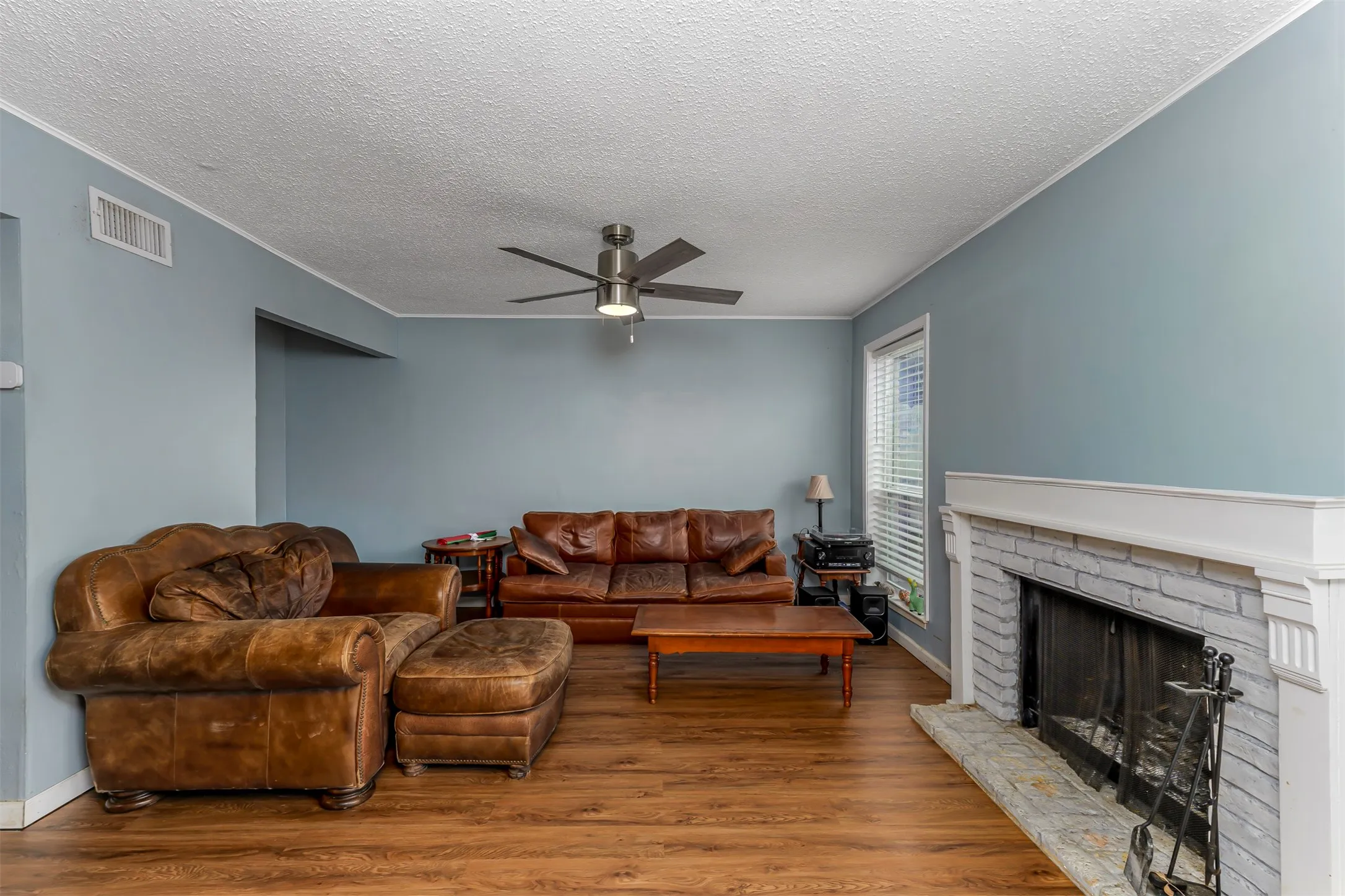 Wood-finish flooring, ceiling fan with integrated lighting, window with blinds, painted brick fireplace with stone hearth, and white mantel