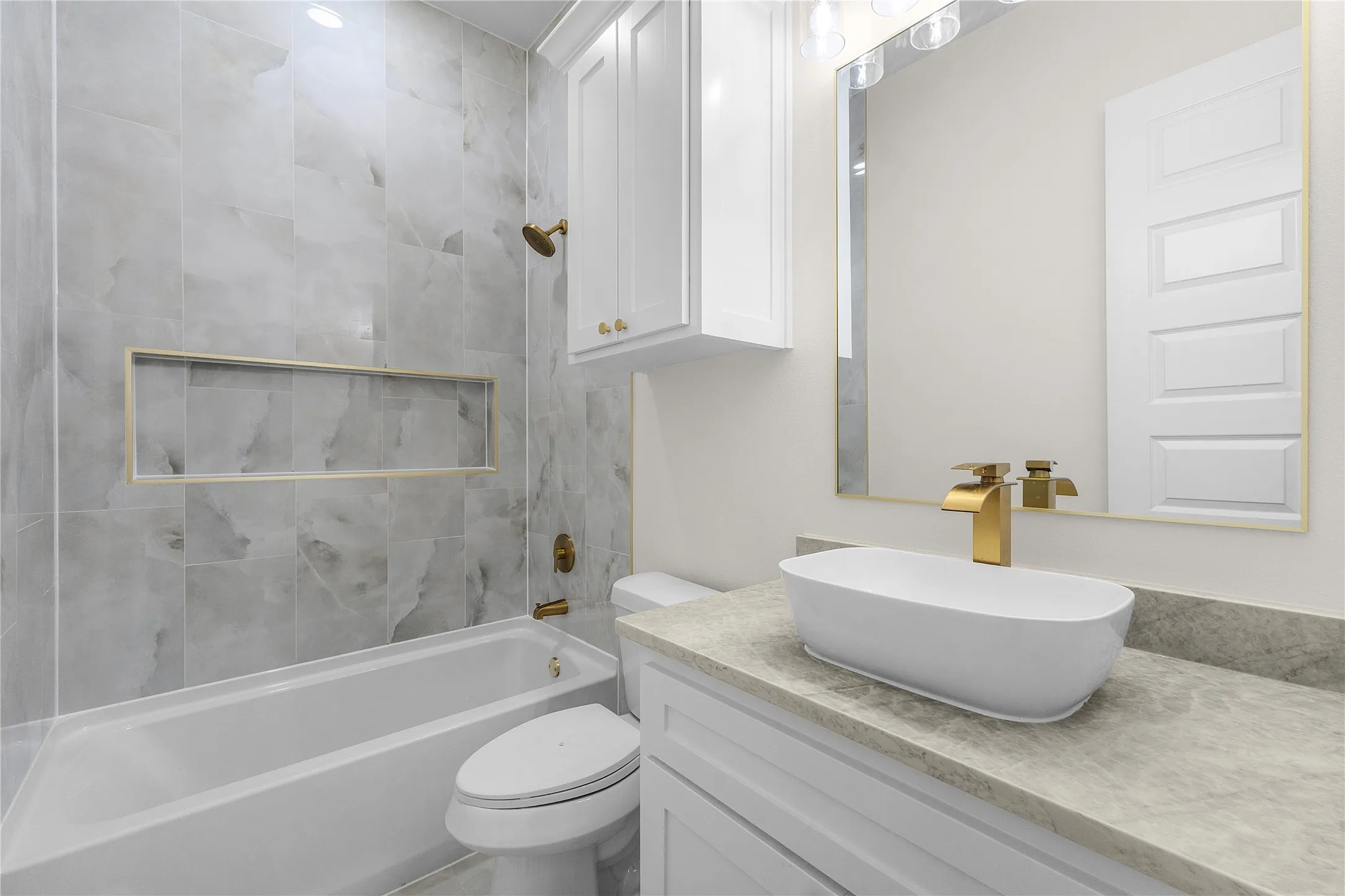 Bathroom featuring a bathtub, tile surround with integrated niche, white vanity, vessel sink with gold-finish faucet, and a large wall mirror