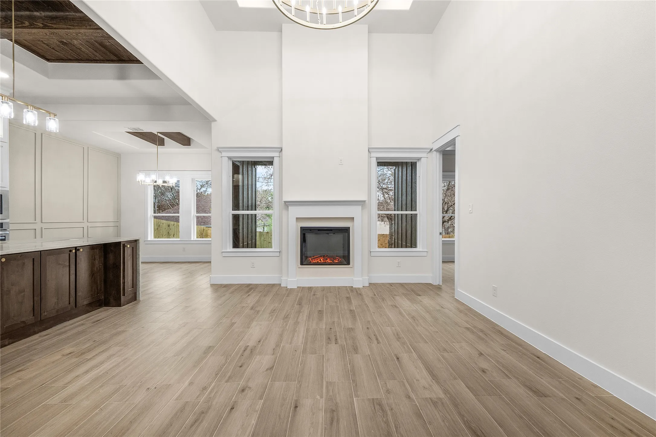Soaring ceilings with a decorative chandelier, white fireplace with mantle, wood-finish flooring, and a kitchen island with dark cabinetry