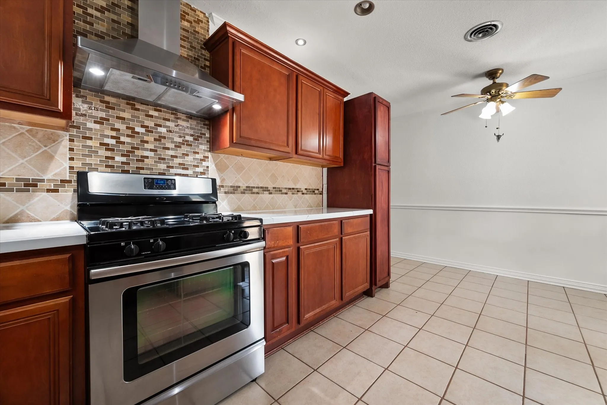 Wood-finish cabinetry, stainless steel range with overhead hood, mosaic tile backsplash, light-toned countertops, and tile flooring