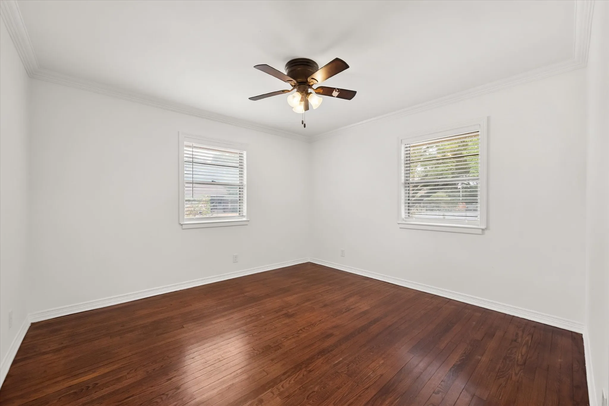 Hardwood flooring, two windows with blinds, ceiling fan with integrated lighting, white walls, and crown molding
