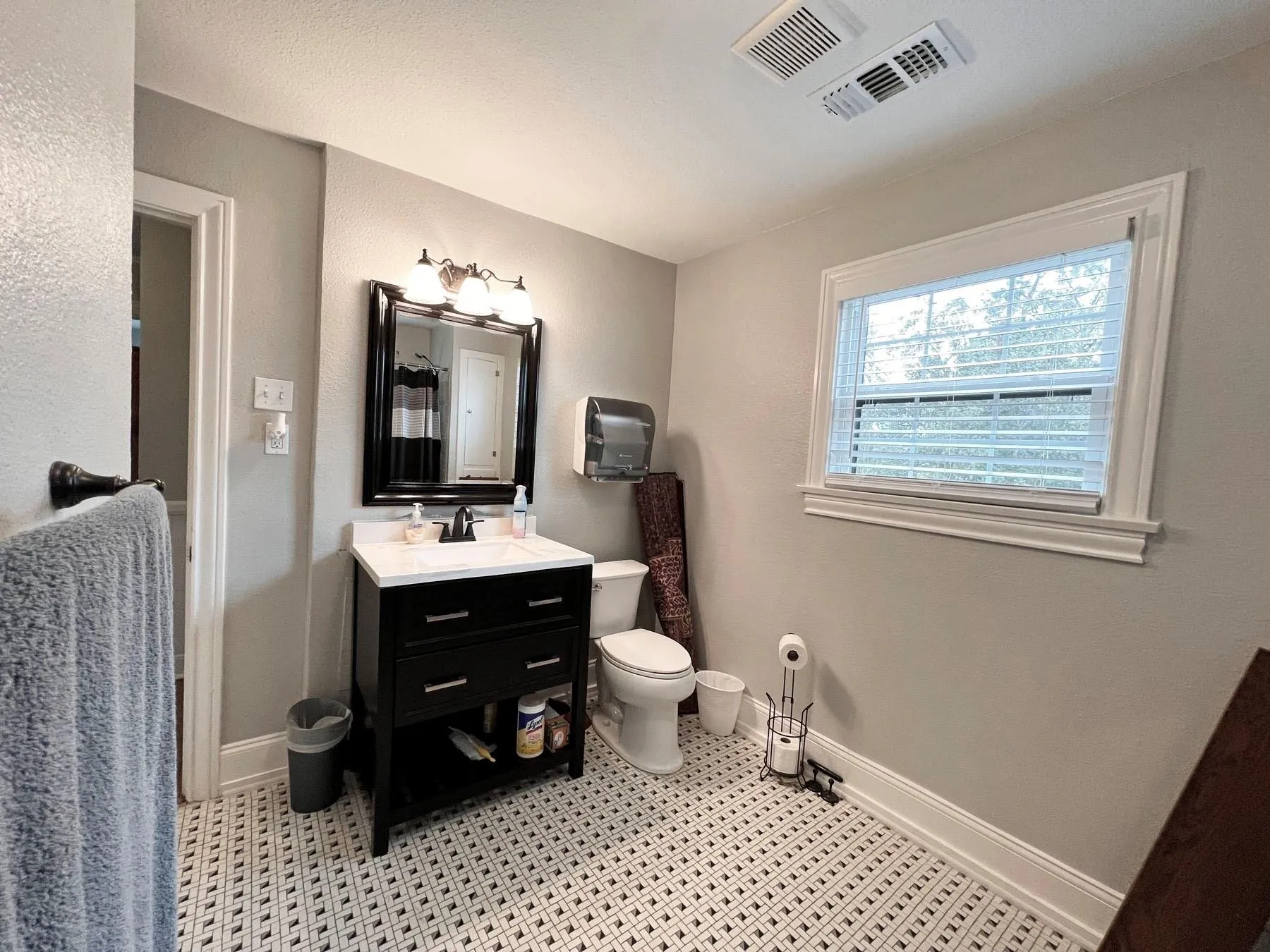White and black patterned tile flooring, black vanity with white countertop, framed vanity mirror with sconce lighting, white window with blinds, and white toilet