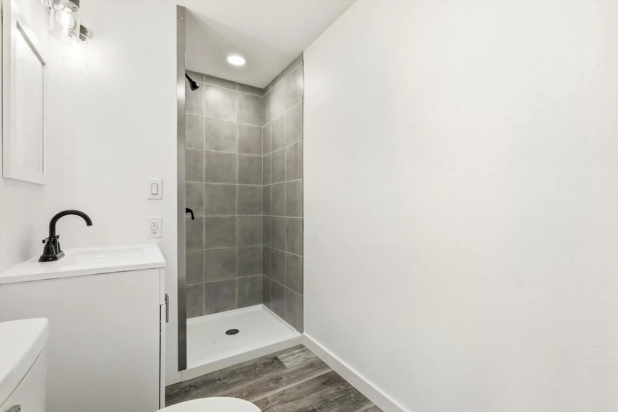 Recessed lighting, wood-finish flooring, white vanity with integrated sink, black matte faucet, and tiled shower surround