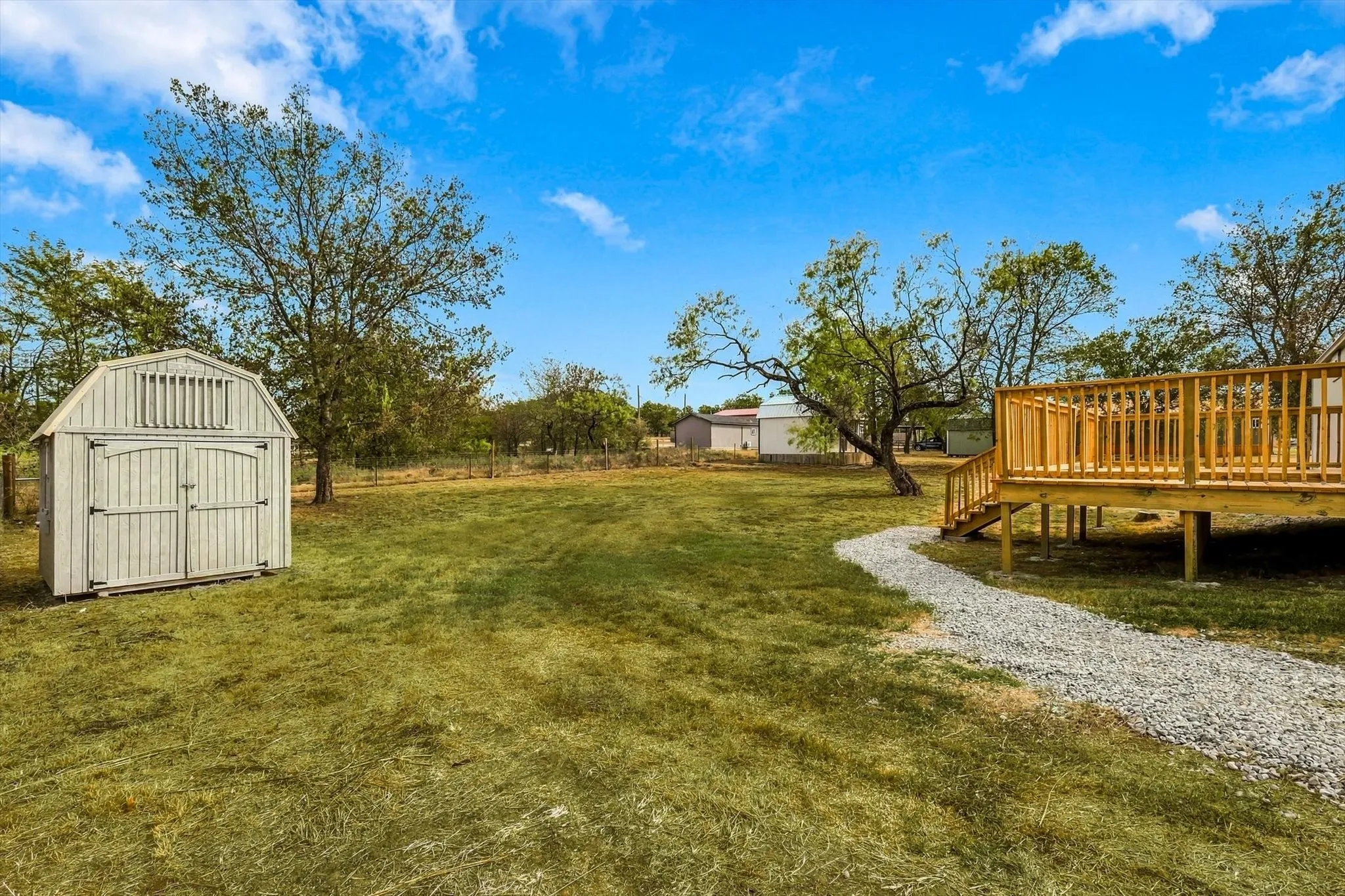 Expansive grassy area featuring a gabled storage shed, gravel pathway, and elevated wood deck