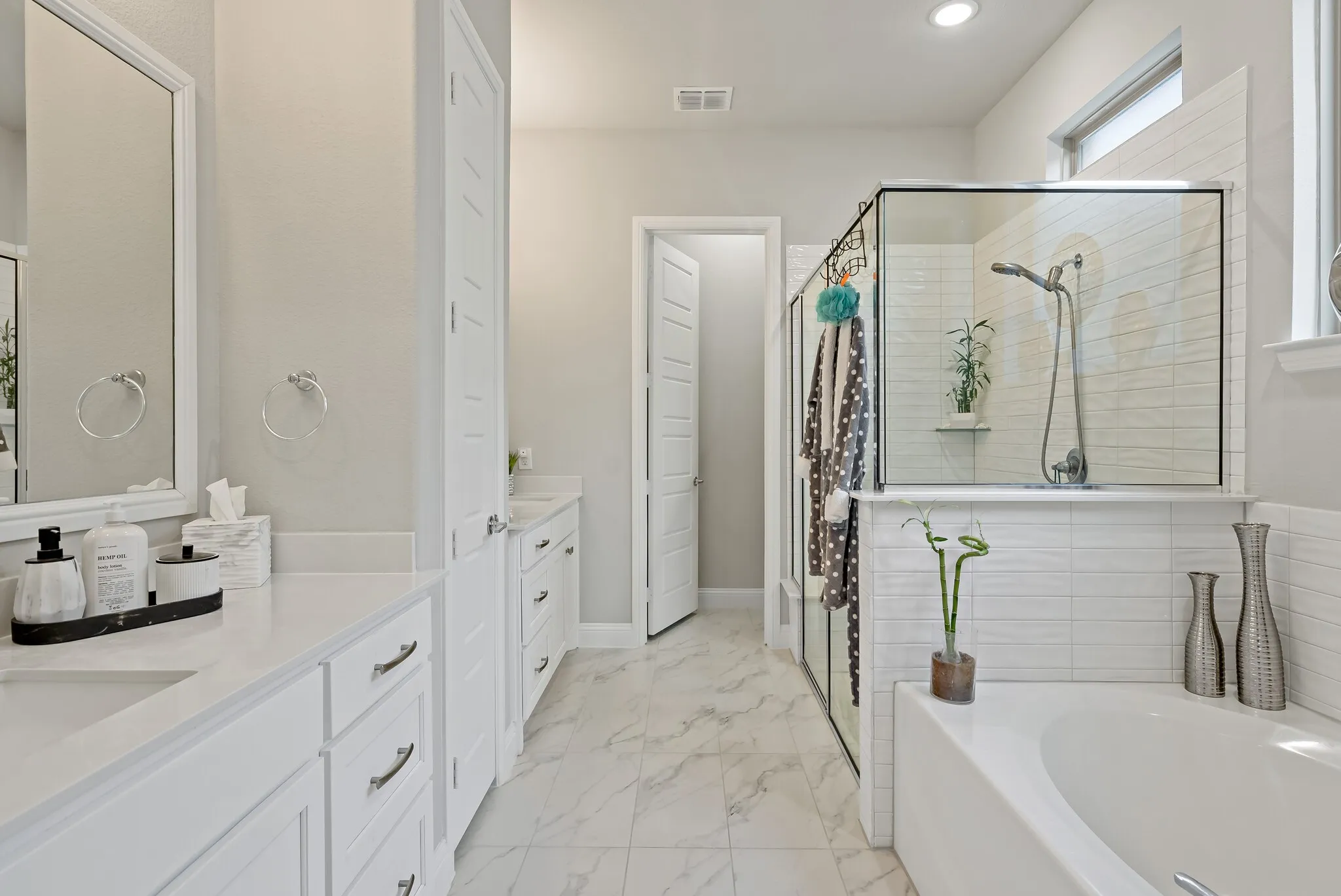 Spacious bathroom featuring a built-in soaking tub, glass-enclosed shower, dual vanities with white cabinetry, and marble-look tile flooring