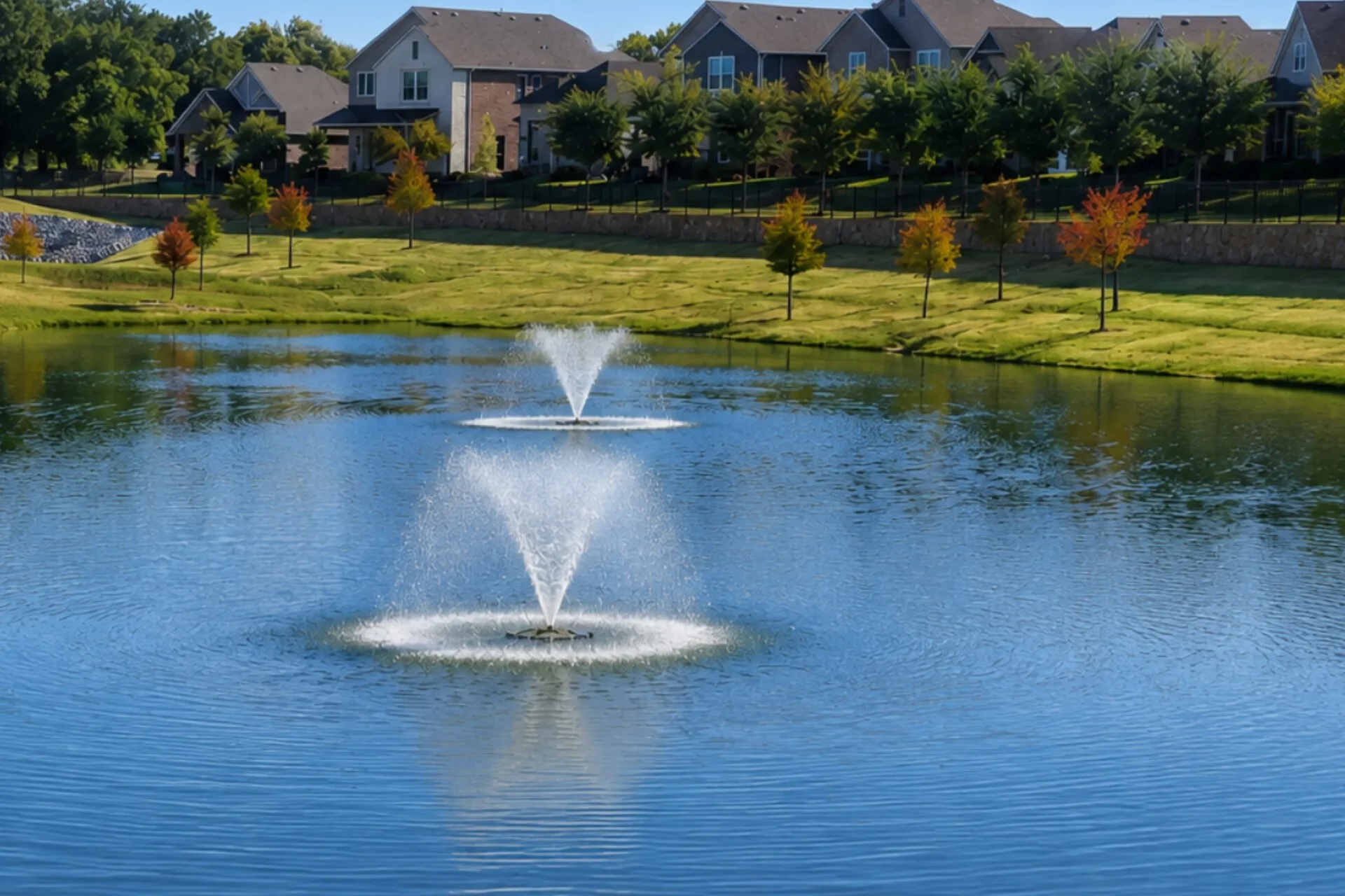 Serene lakefront setting featuring manicured grounds with mature landscaping, dual decorative fountains, and expansive open lawn bordered by residential architecture and tree-lined perimeter