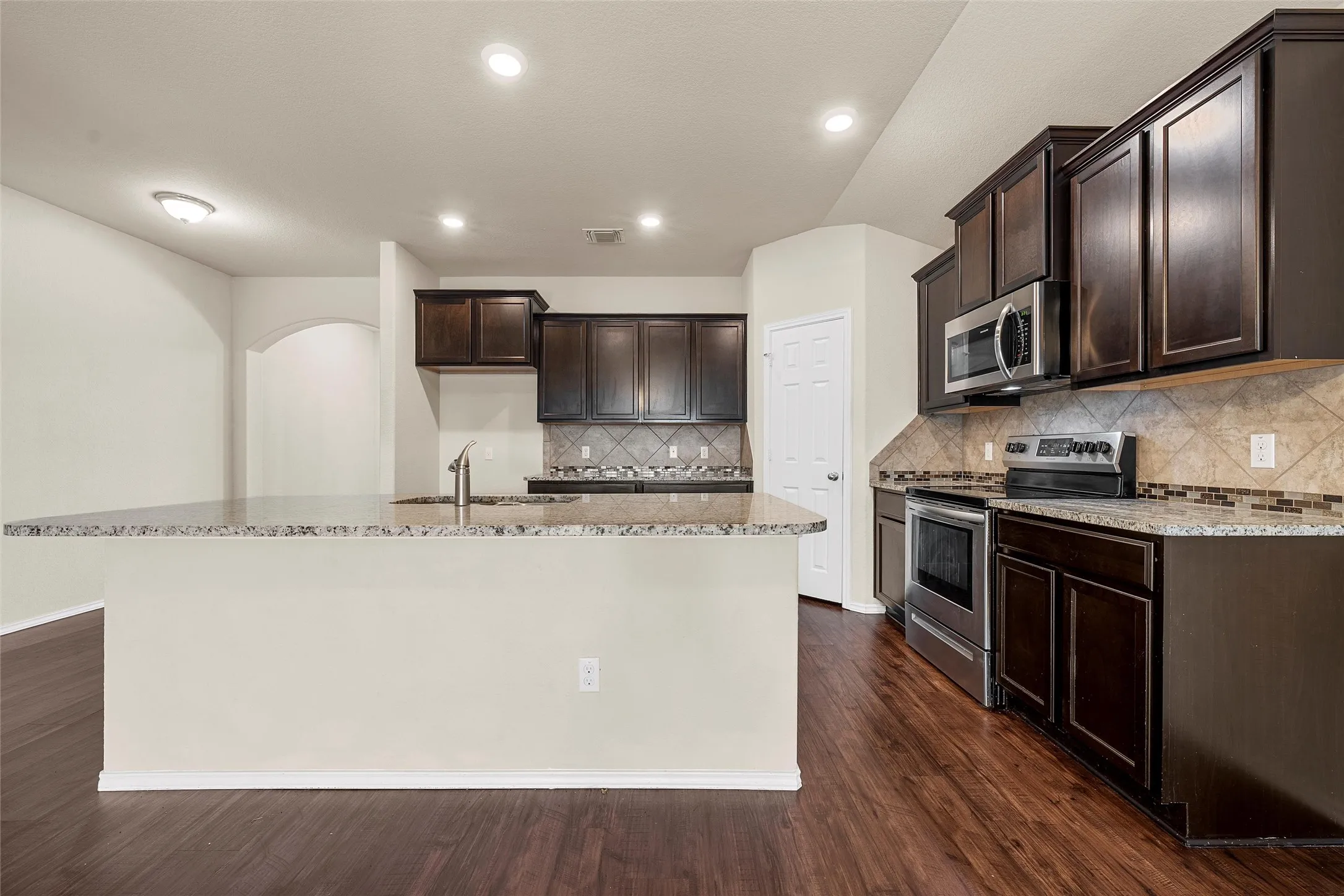 Kitchen with appliances with stainless steel finishes, light stone counters, dark brown cabinets, an island with sink, and dark wood-style floors