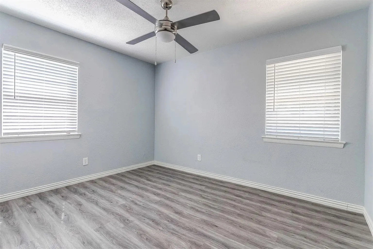 Wood-finish flooring, two windows with blinds, and a ceiling fan with integrated lighting