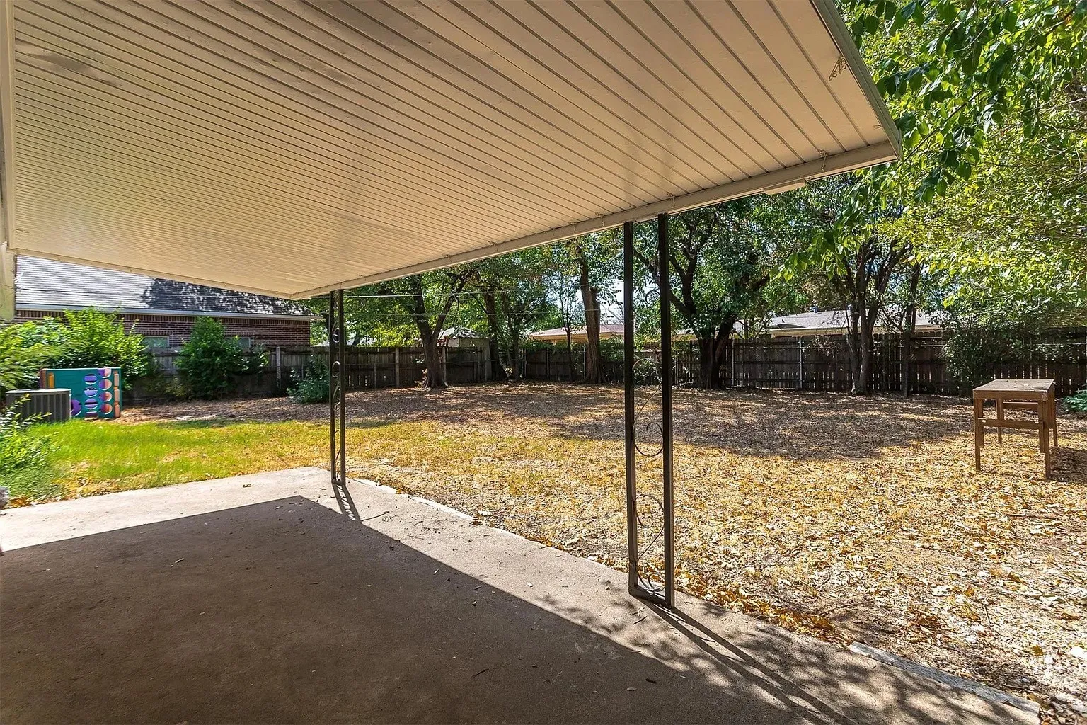 Covered patio with a concrete slab, leading to an expansive yard with mature trees and a wood fence