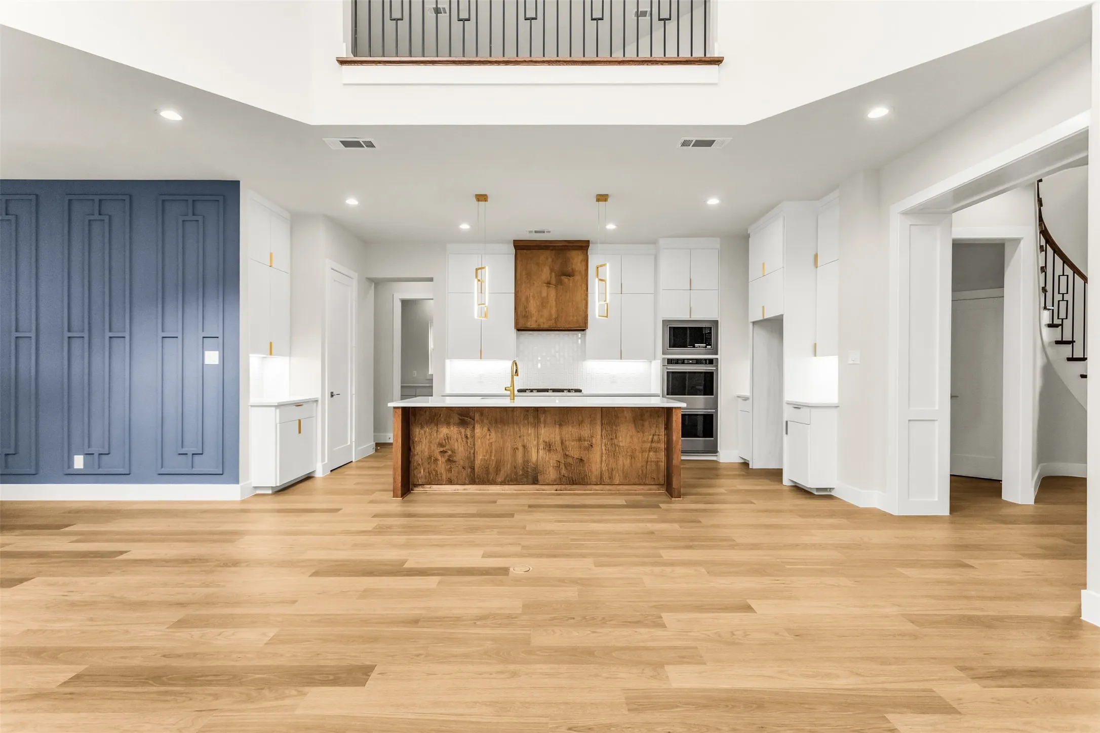 Open-concept interior featuring wood-finish flooring, a kitchen island with stone countertop, built-in double ovens, and a decorative accent wall