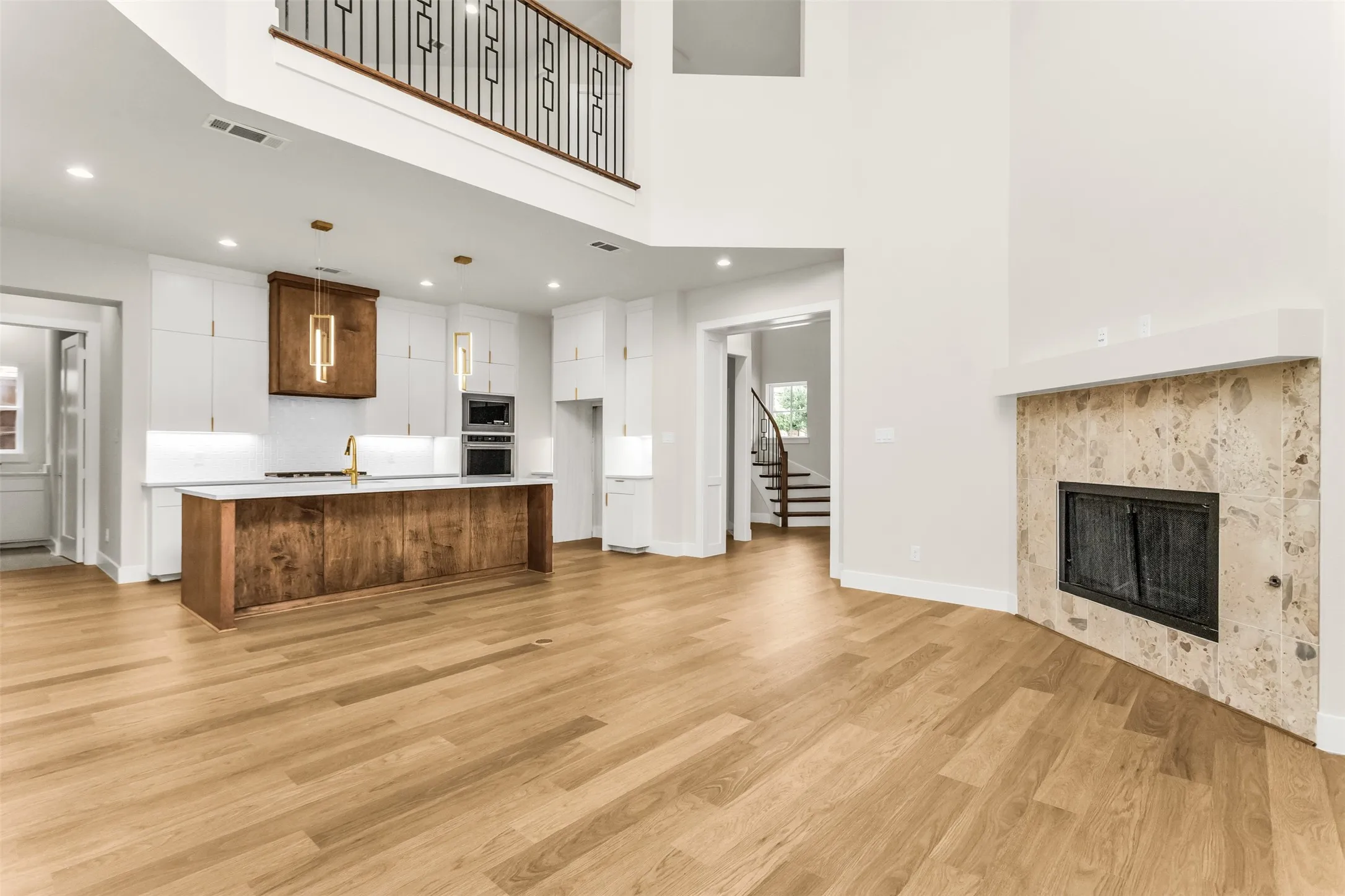 Vaulted ceilings, a kitchen island with built-in appliances, wood-finish flooring, a stone-clad fireplace, and an upper-level balcony with a wrought iron railing