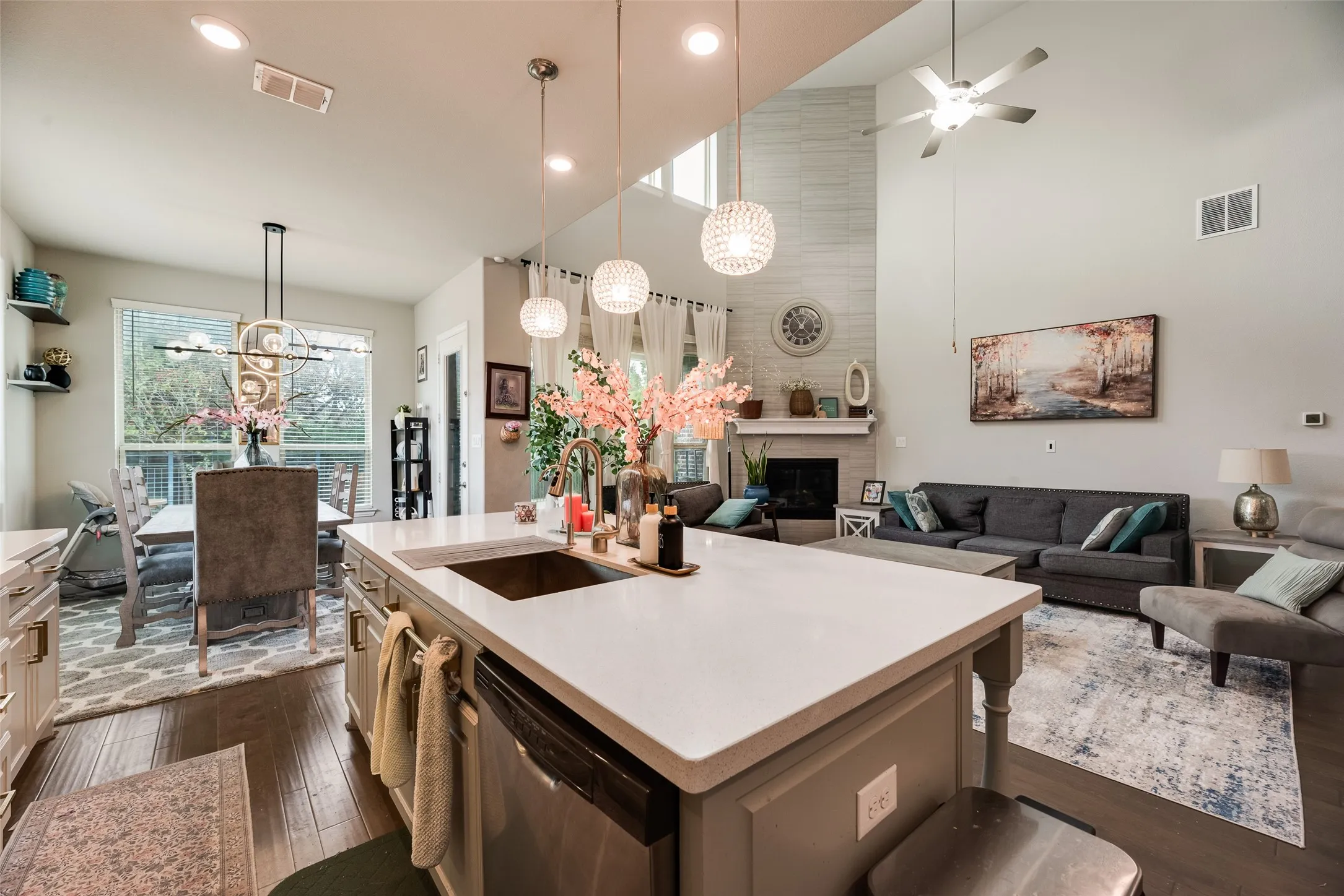 Kitchen island with an undermount sink and three pendant lights, wood-finish flooring, a dining area with a linear chandelier, a living area with a fireplace, and a high vaulted ceiling
