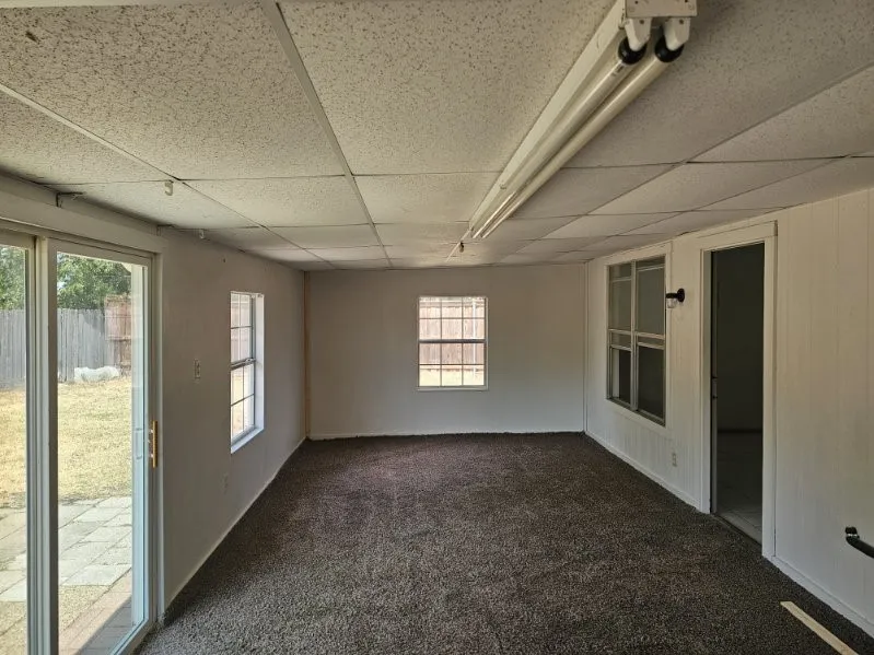Carpeted flooring, multiple windows, sliding glass door, drop ceiling with fluorescent lighting, and a white paneled wall