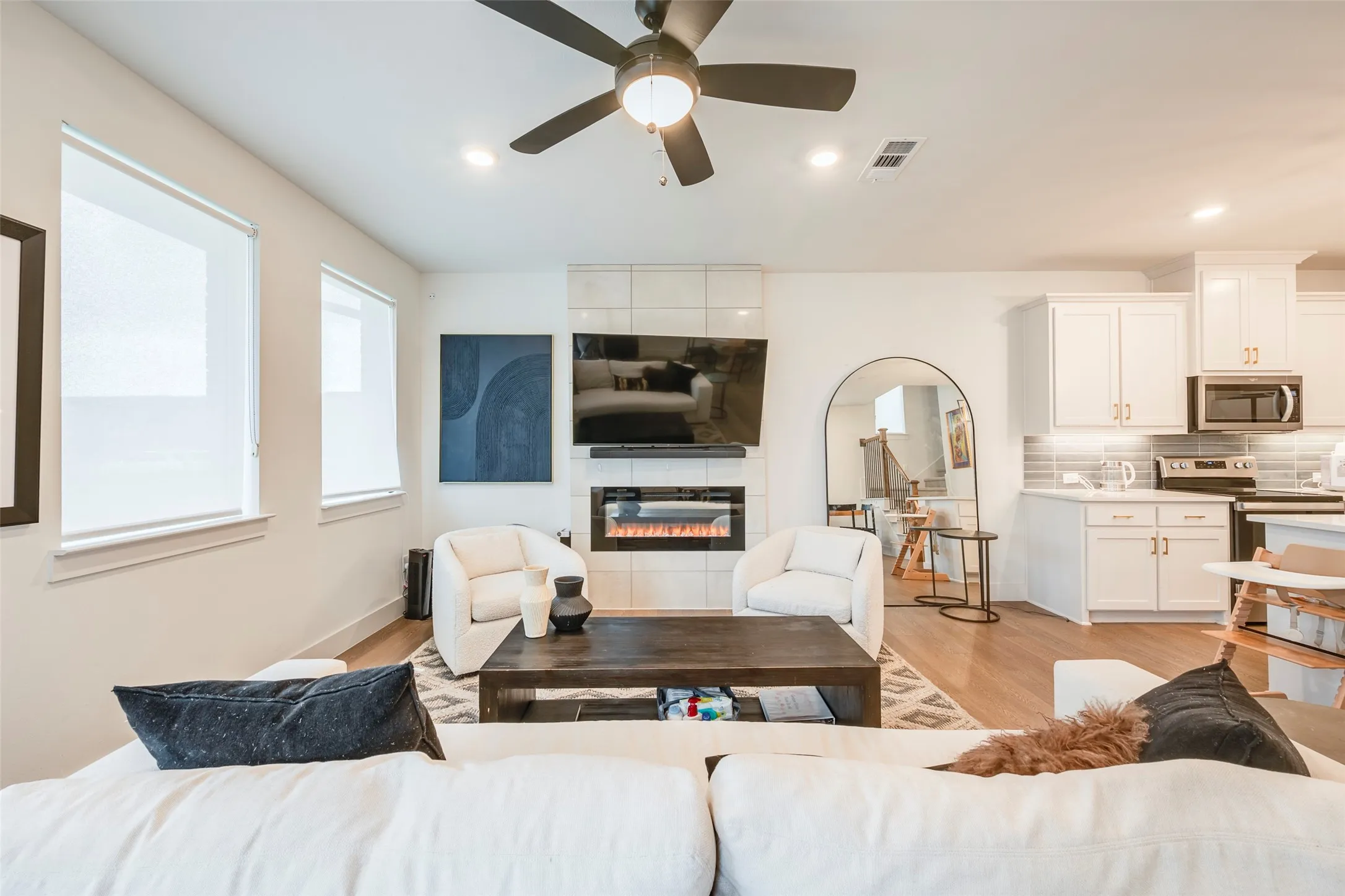 Open-concept living area featuring wood-finish flooring, a contemporary fireplace with tile surround, and an integrated kitchen with white cabinetry and stainless steel appliances