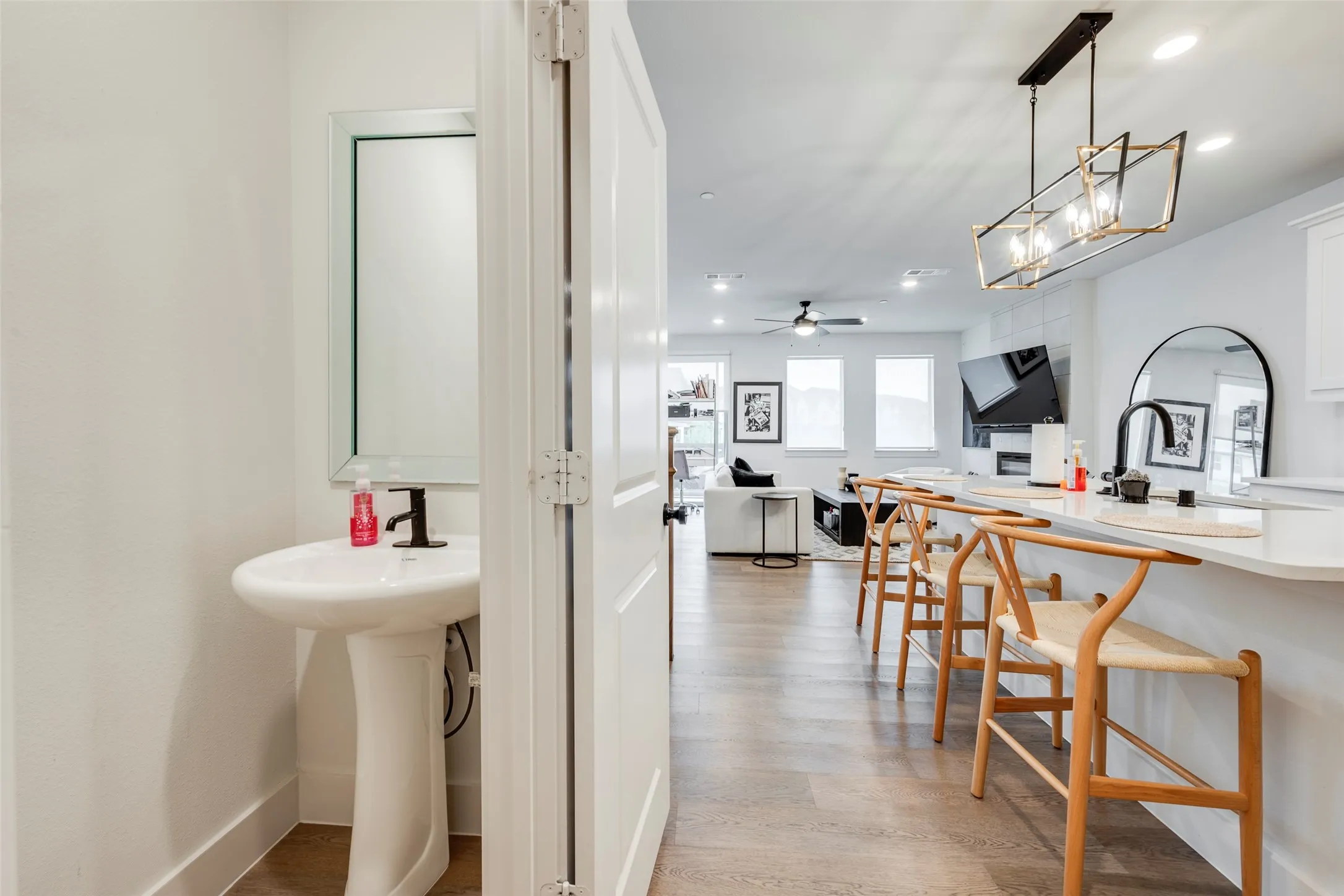 Pedestal sink with black faucet, wood-finish flooring, white kitchen island with countertop seating, geometric pendant lighting fixture, and a ceiling fan