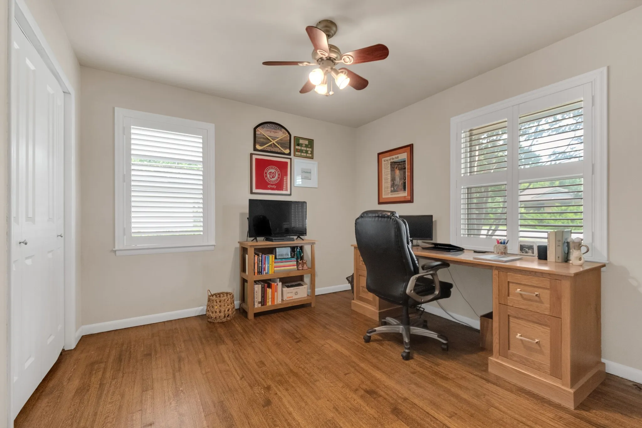 Room featuring wood-finish flooring, two windows with white plantation shutters, and a ceiling fan with integrated lighting