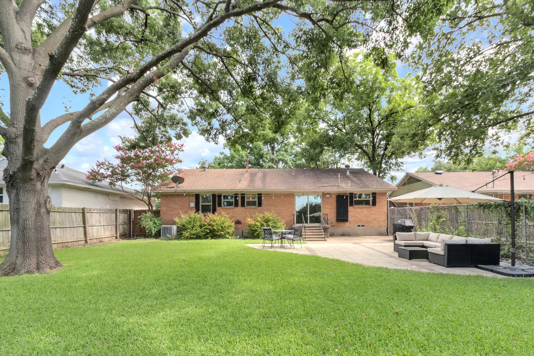Expansive backyard featuring a manicured lawn, large shade tree, brick exterior, full privacy fencing, and a concrete patio