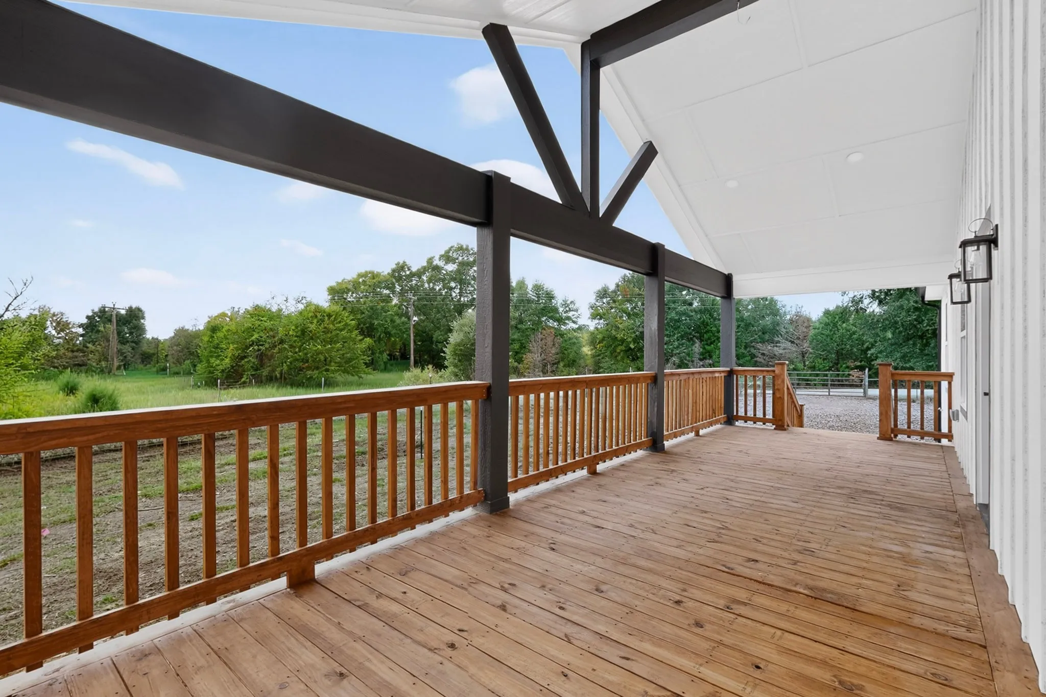 Spacious covered porch with wood-finish decking and railing, supported by dark-stained structural beams, featuring a white vaulted ceiling and exterior wall sconces