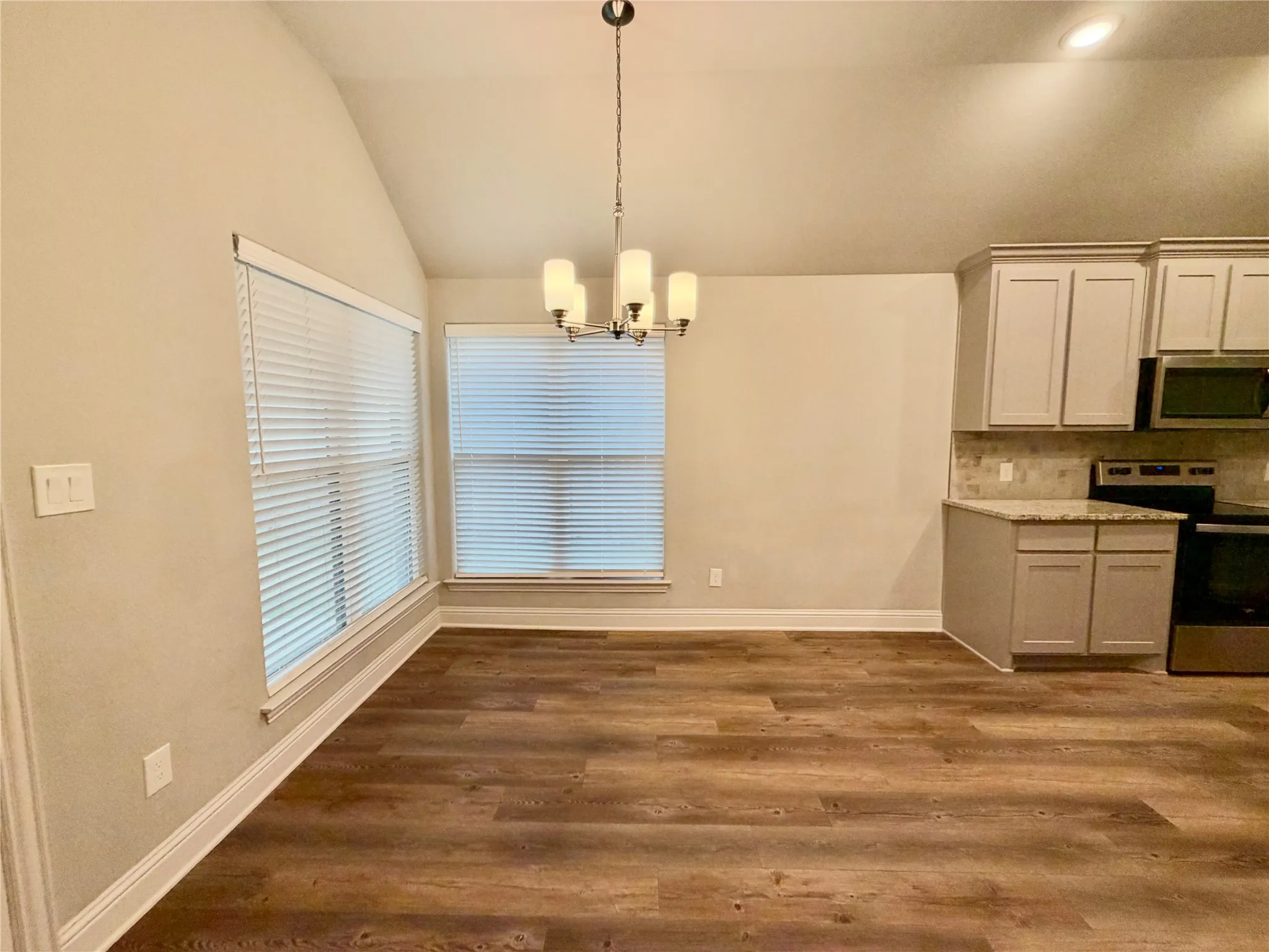 Vaulted ceiling with pendant lighting, wood-finish flooring, two windows with blinds, built-in cabinetry with light-colored countertops, and tiled backsplash