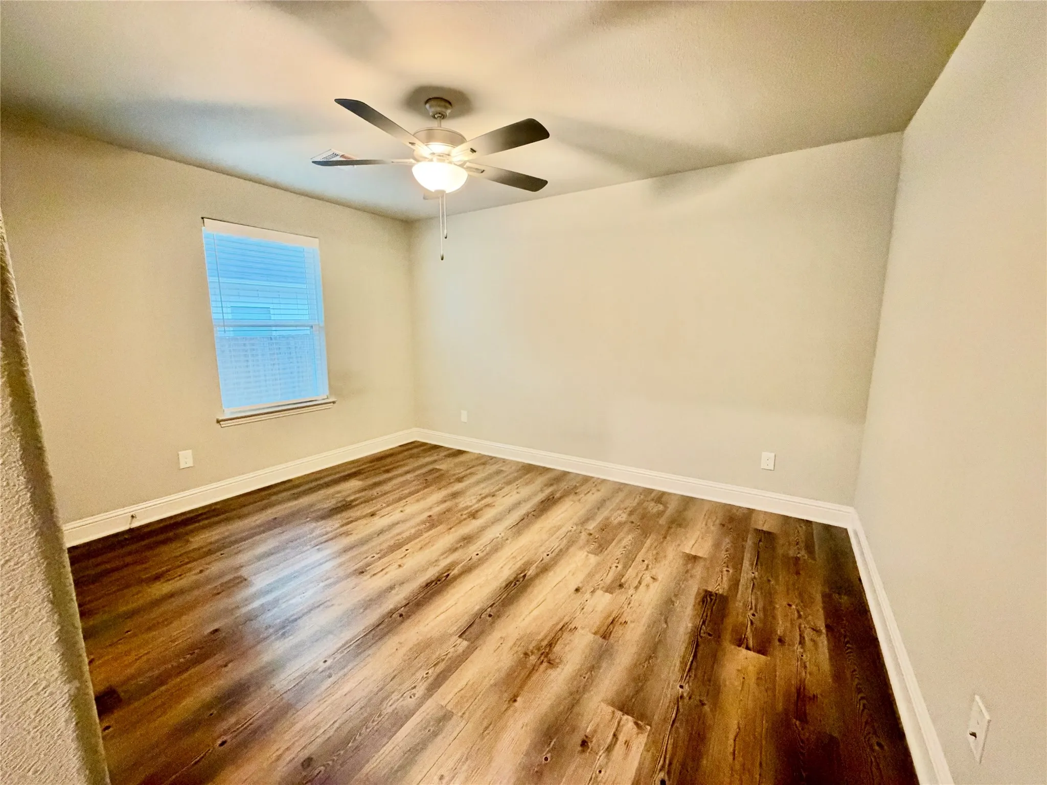 Wood-finish flooring with white trim, light neutral wall paint, single window with blinds, and a ceiling fan with integrated lighting