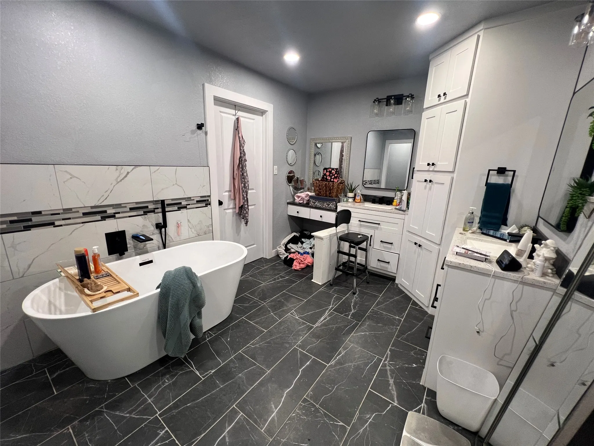 Freestanding soaking tub, black-veined tile flooring, white shaker cabinetry, white countertop, and tiled accent wall
