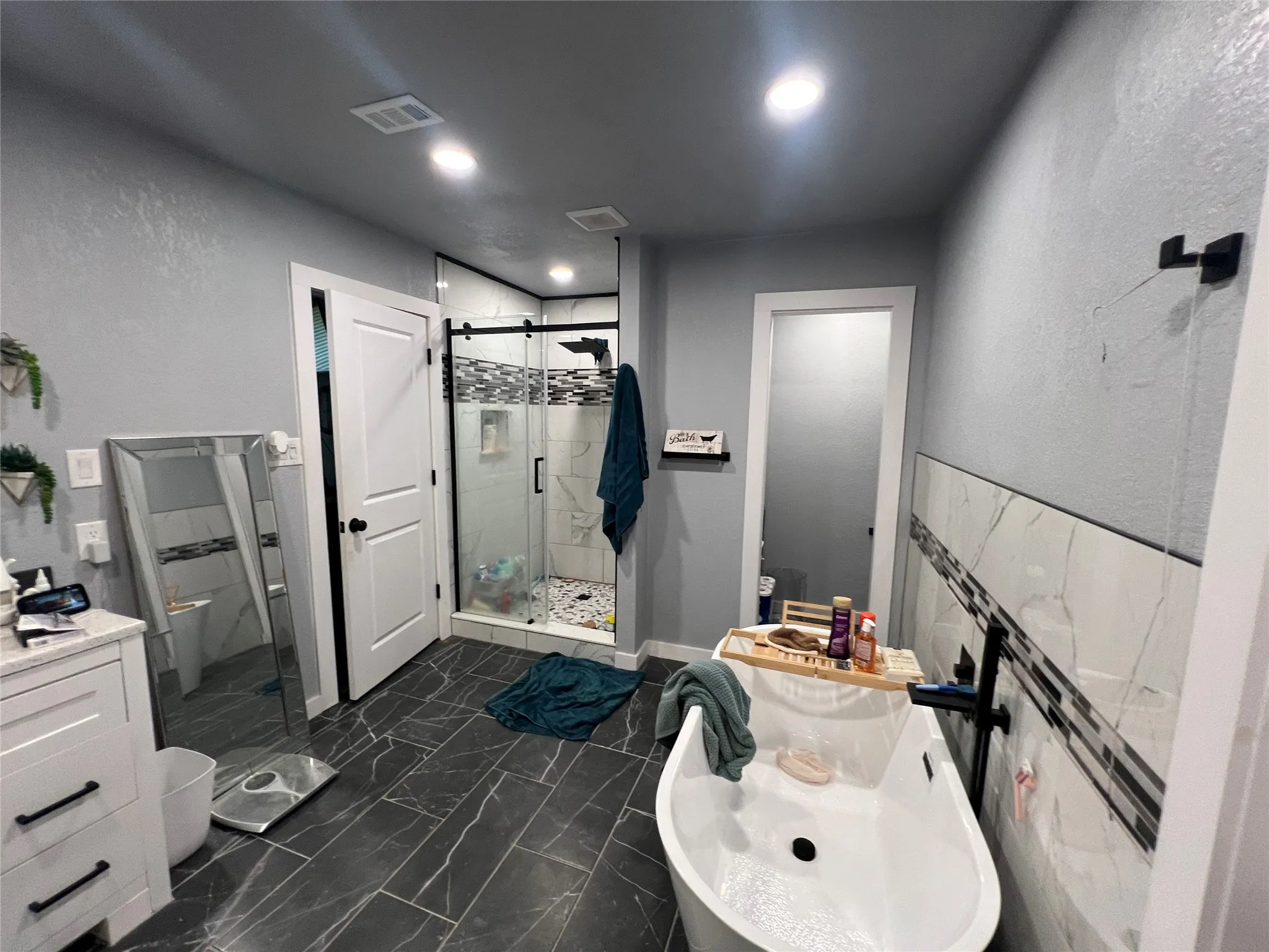 Freestanding soaking tub with a wall-mounted faucet, walk-in shower with a glass enclosure and sliding door, and dark-toned floor tiles with light veining