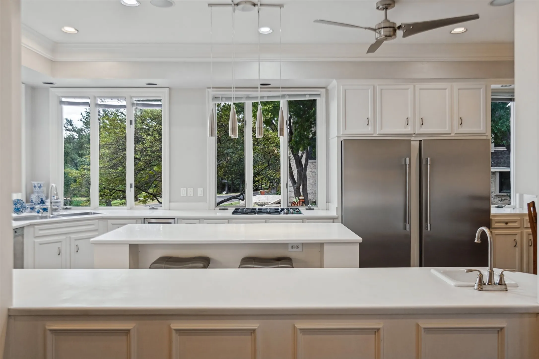 Chef's kitchen featuring stainless steel refrigerator, quartz countertops, white cabinetry with paneled doors, large center island with seating, and abundant natural light from expansive multi-pane windows overlooking mature landscaping
