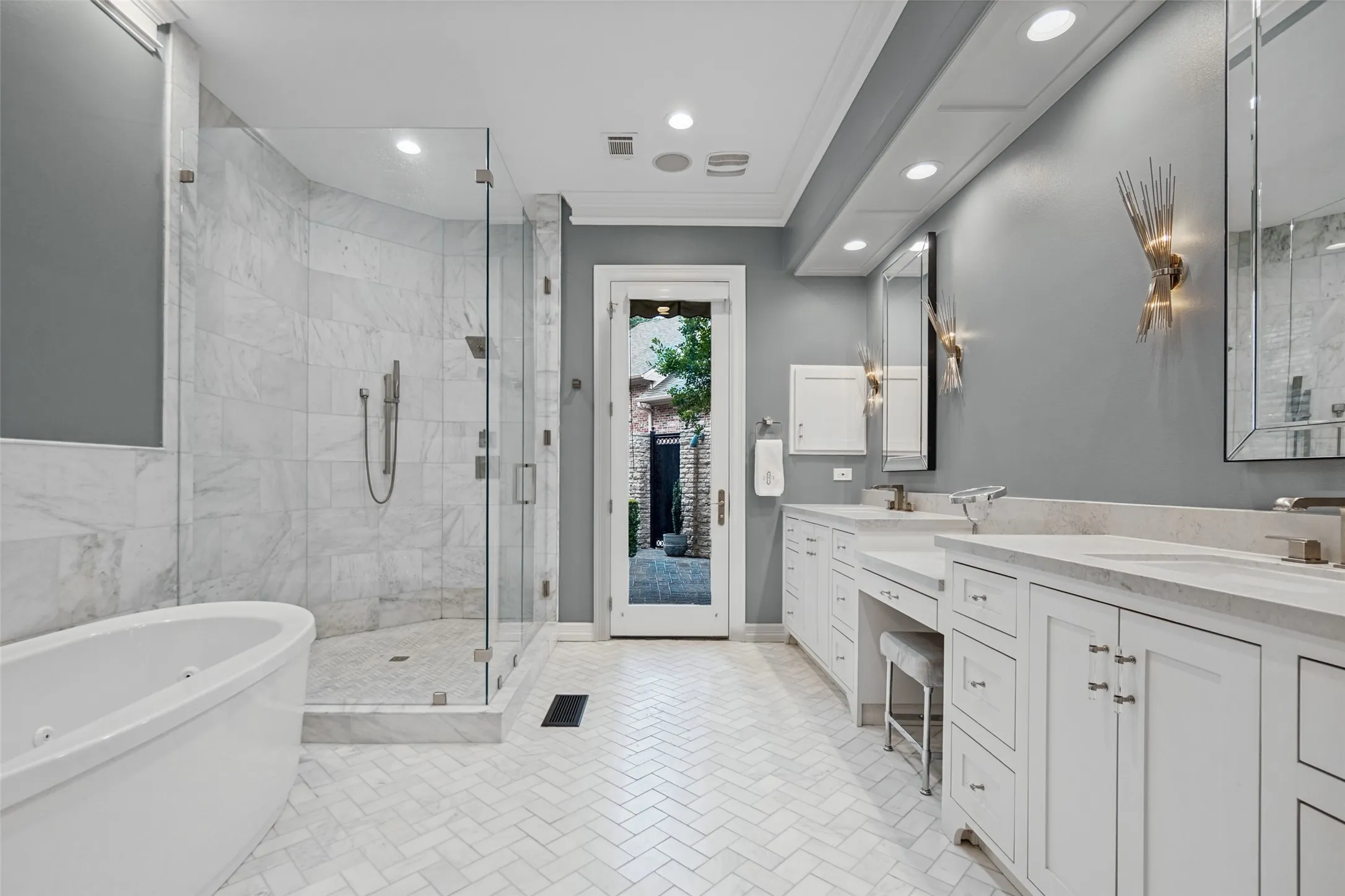 Freestanding soaking tub, walk-in shower with glass enclosure, dual vanity with undermount sinks, white cabinetry, and herringbone tile flooring
