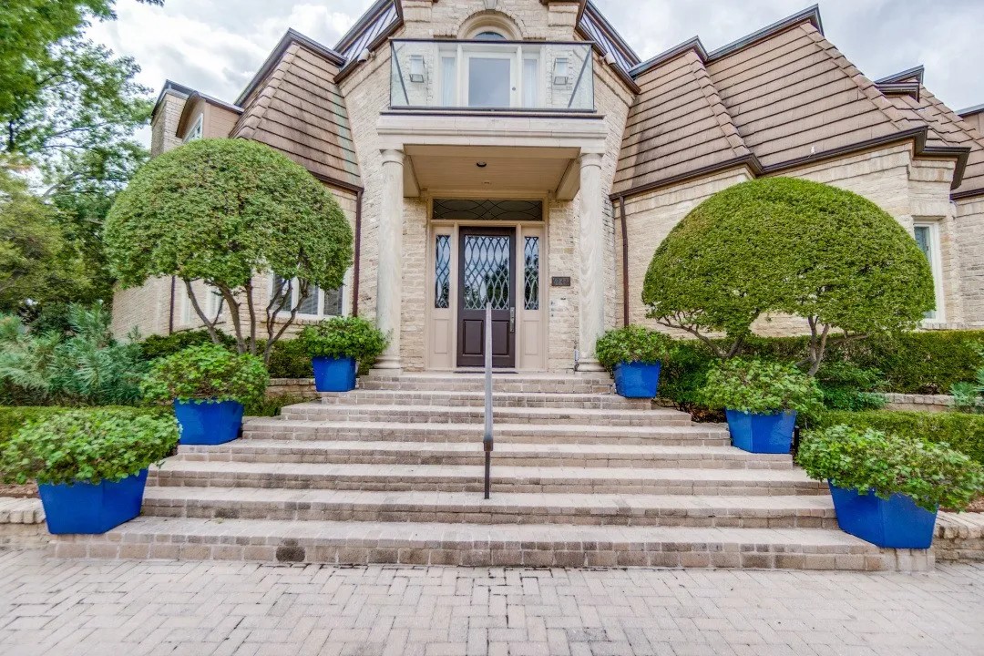 Light-toned brick exterior with a prominent entrance featuring a dark wood door, stone steps, and a glass-paneled balcony