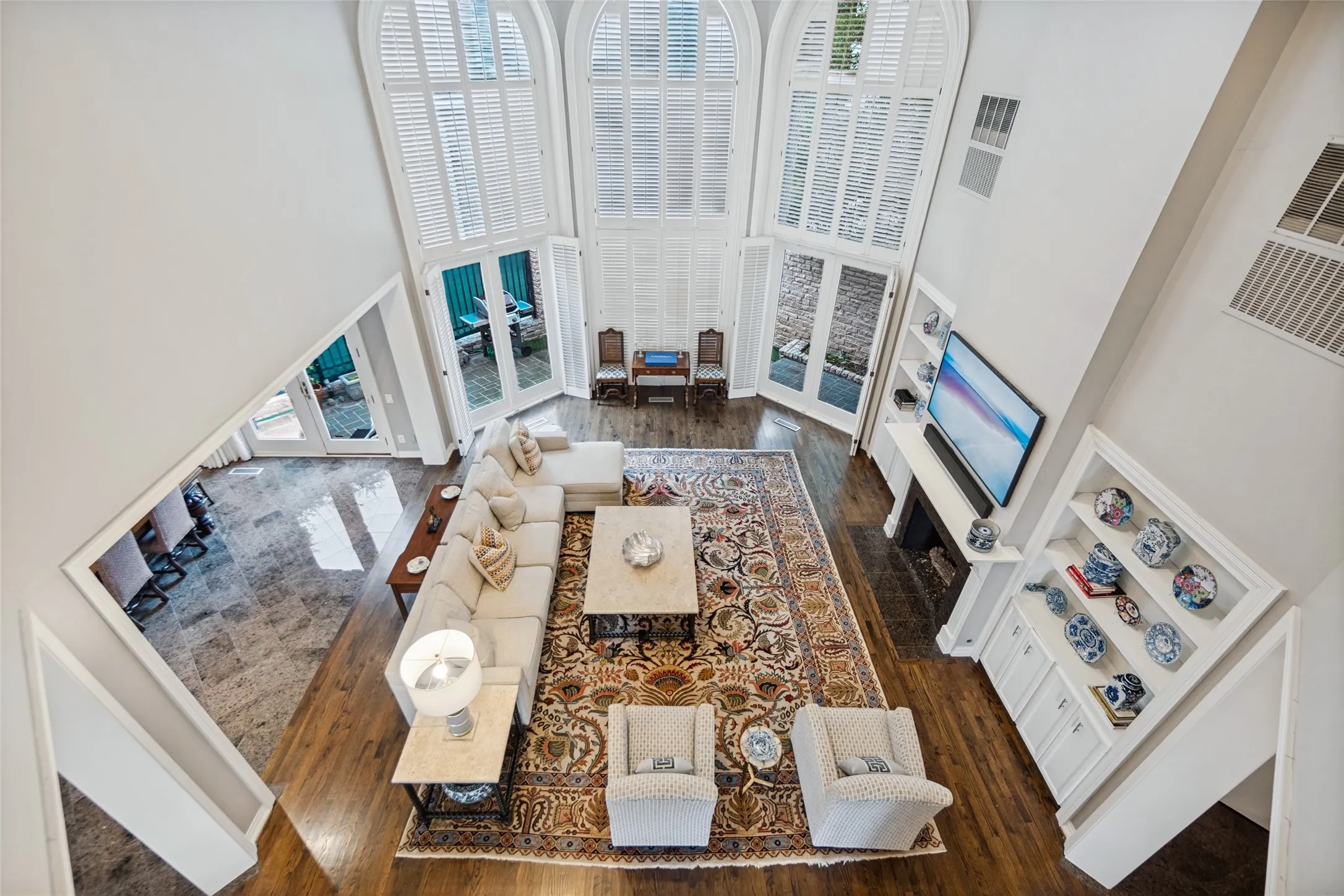 Double-height ceilings, arched windows with plantation shutters, rich wood-finish flooring, a built-in fireplace with mantel, and integrated shelving with lower cabinetry