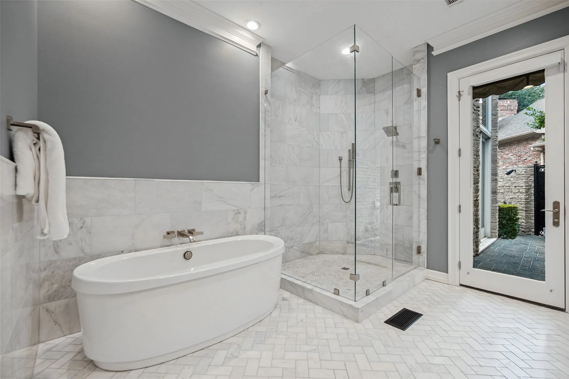Freestanding soaking tub, walk-in shower with clear glass enclosure, marble-style tile walls, herringbone tile flooring, and direct outdoor access