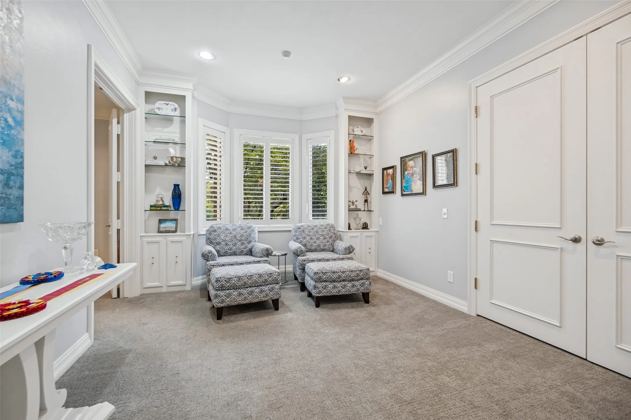Built-in shelving with glass fronts, bay windows with plantation shutters, crown molding, recessed lighting, and carpet flooring