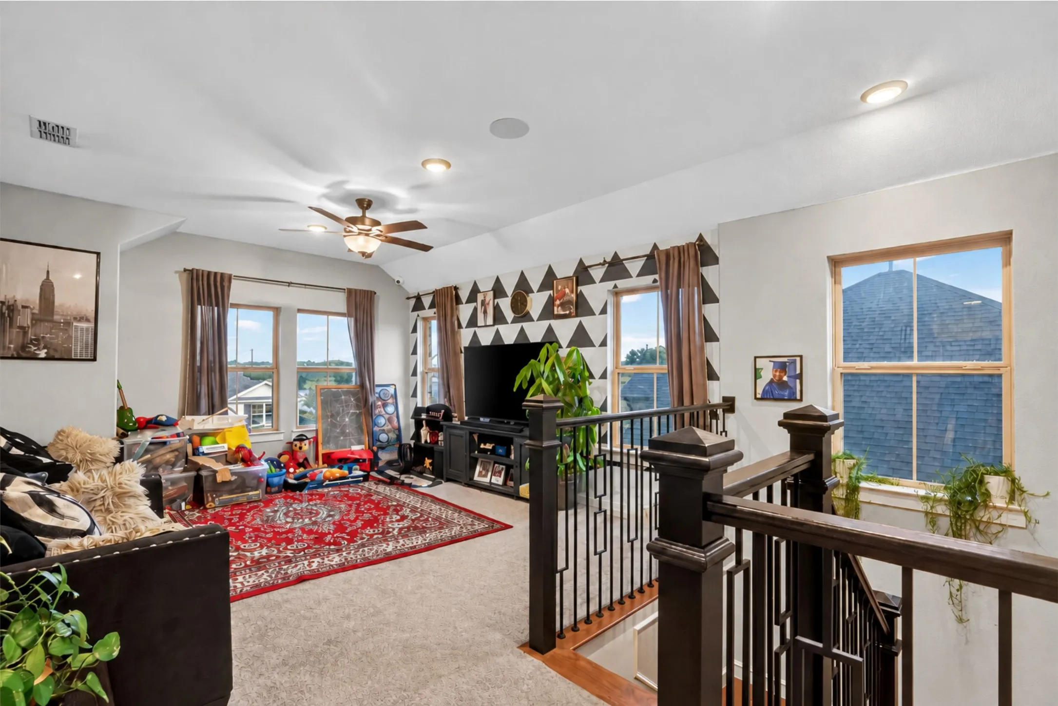 Carpeted living area featuring multiple windows, a ceiling fan with integrated lighting, recessed lighting, and a dark wood and metal railing