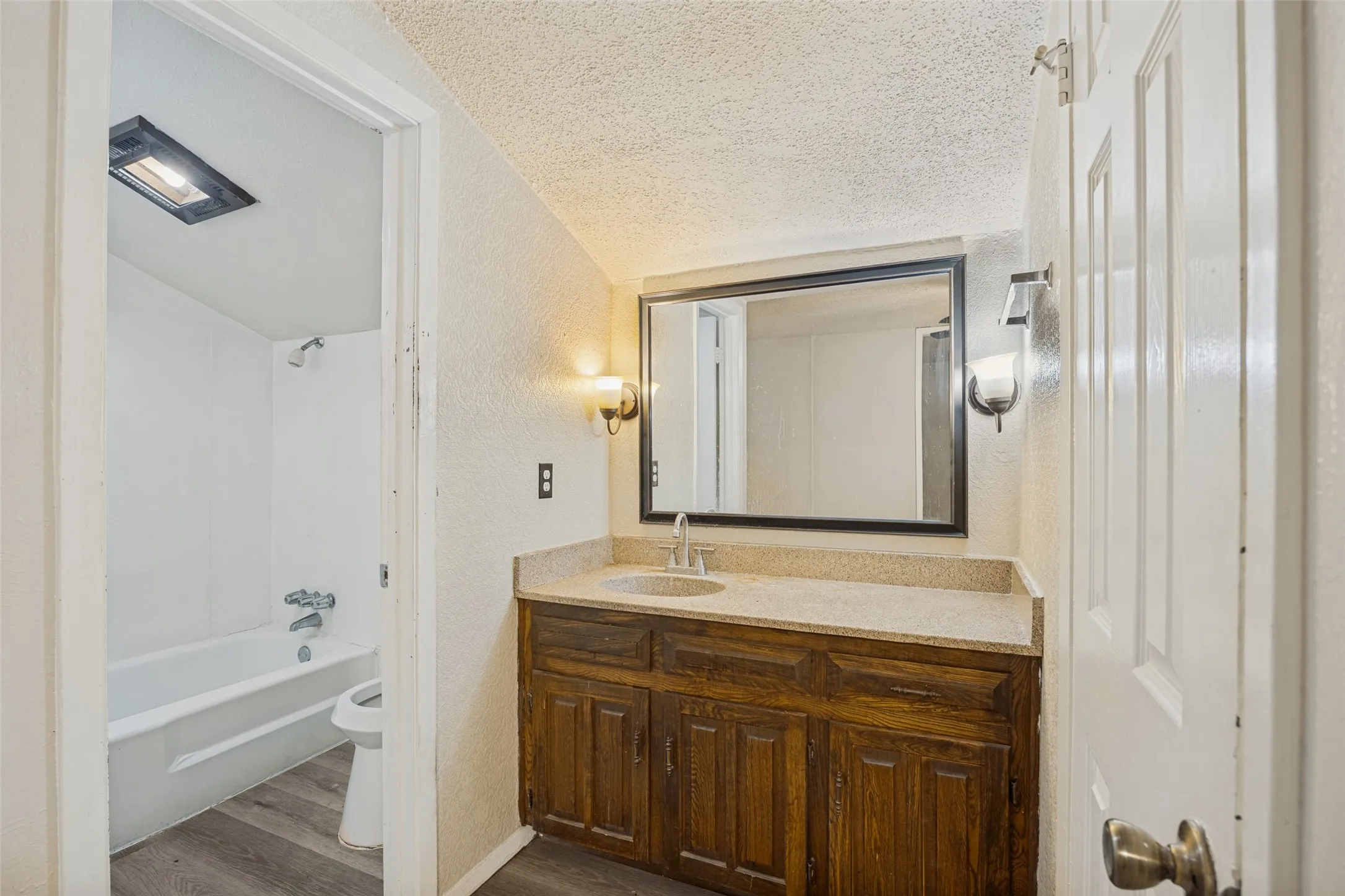 Wood-finish flooring, wood vanity with integrated sink, wall-mounted sconces, and a framed mirror
