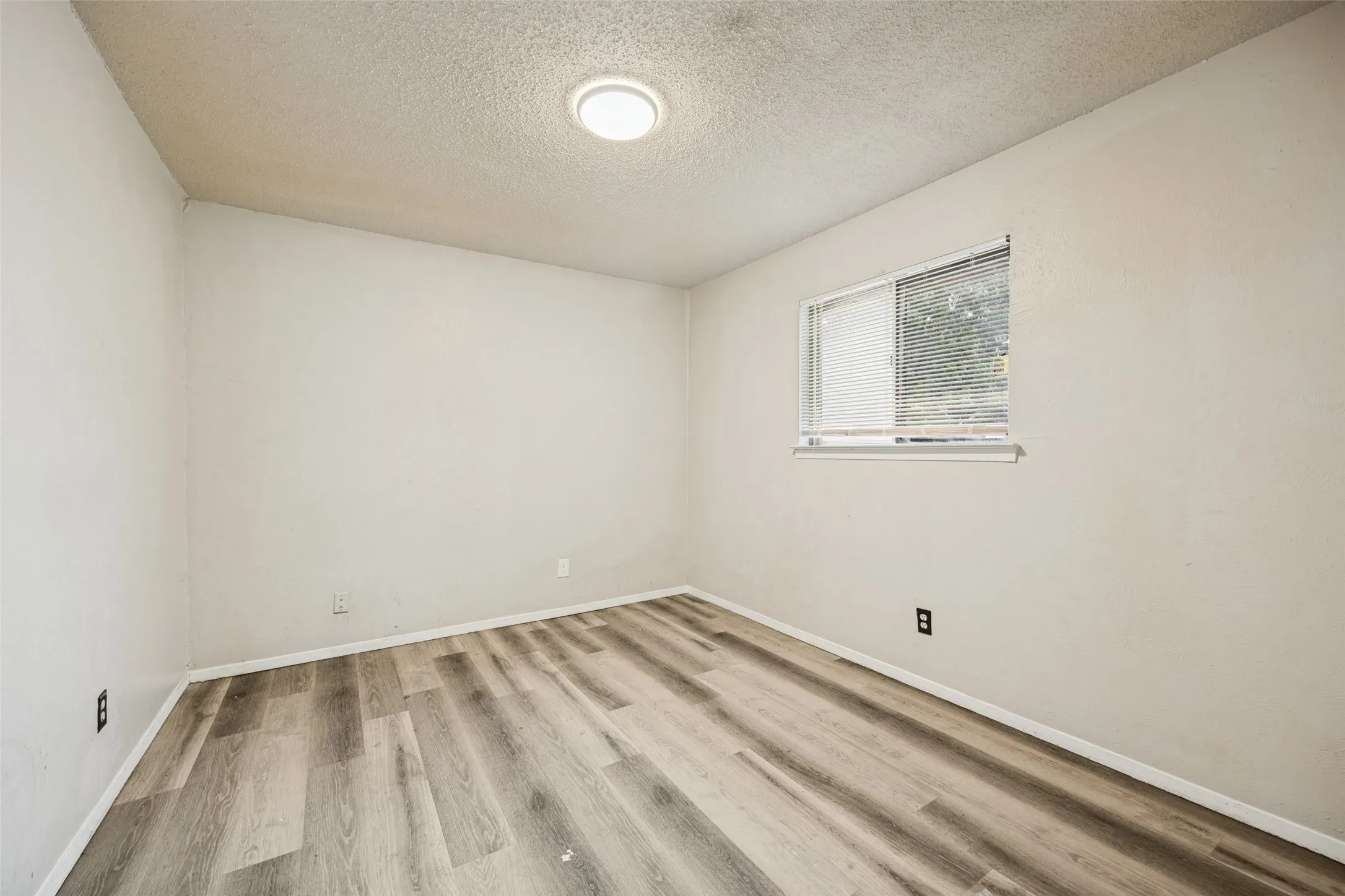 Wood-finish flooring, single window with blinds, overhead flush-mount lighting, and neutral wall finishes