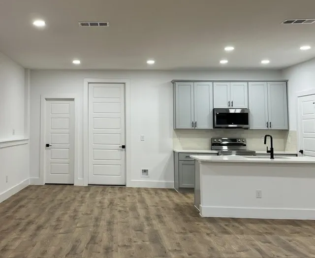 Kitchen area featuring light gray cabinetry, stainless steel appliances, a white island with a matte black faucet, and wood-finish flooring