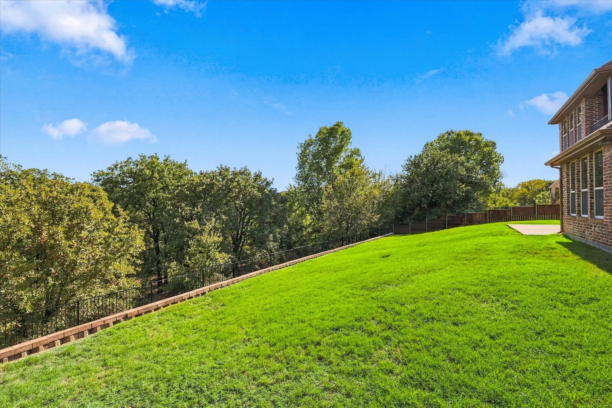Expansive backyard featuring a sloping grass lawn, tiered retaining wall, perimeter fencing, and mature trees