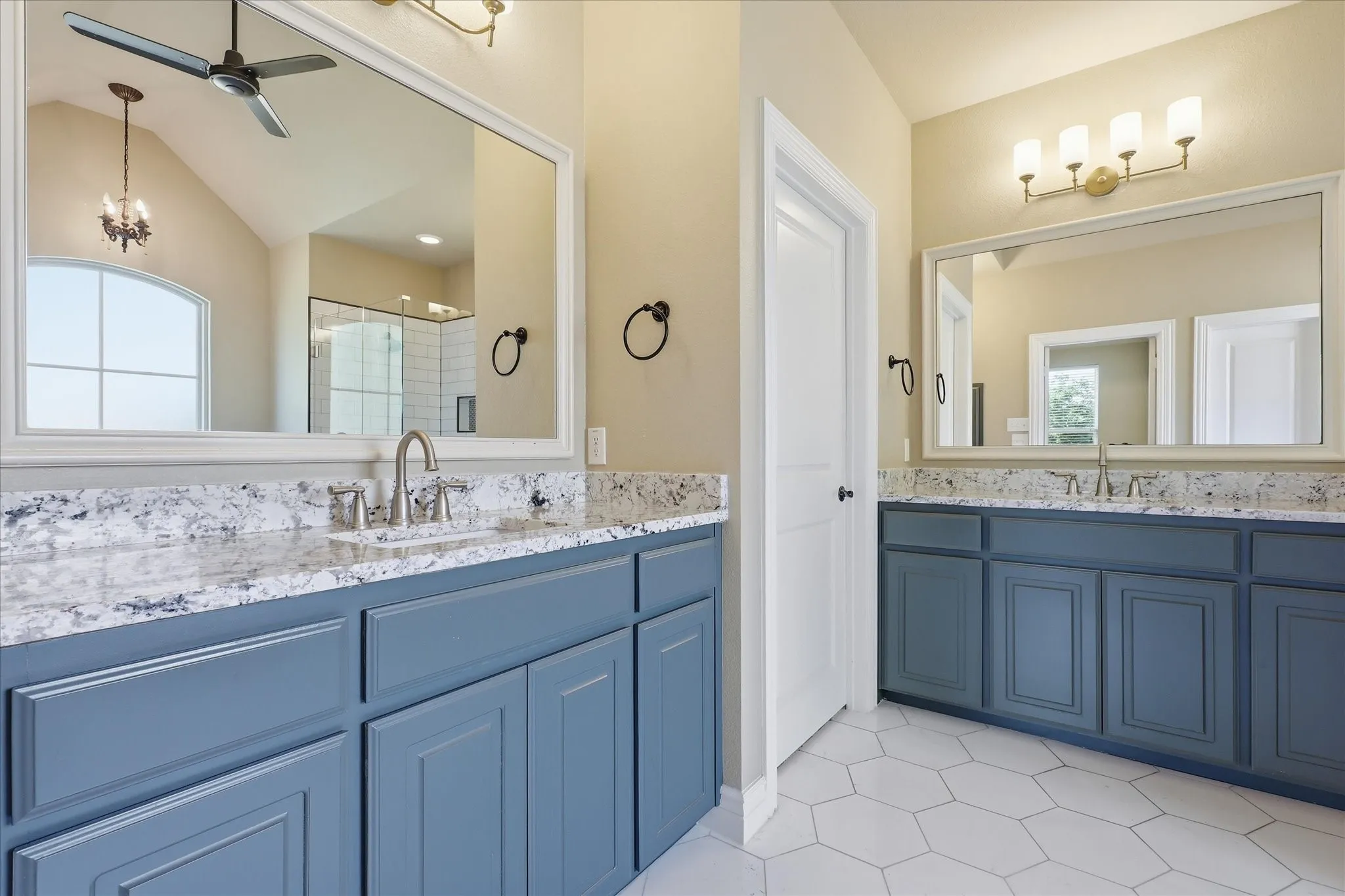 Dual vanity featuring blue cabinetry, patterned stone-finish countertops, rectangular undermount sinks, large framed mirrors, and white hexagonal floor tiling