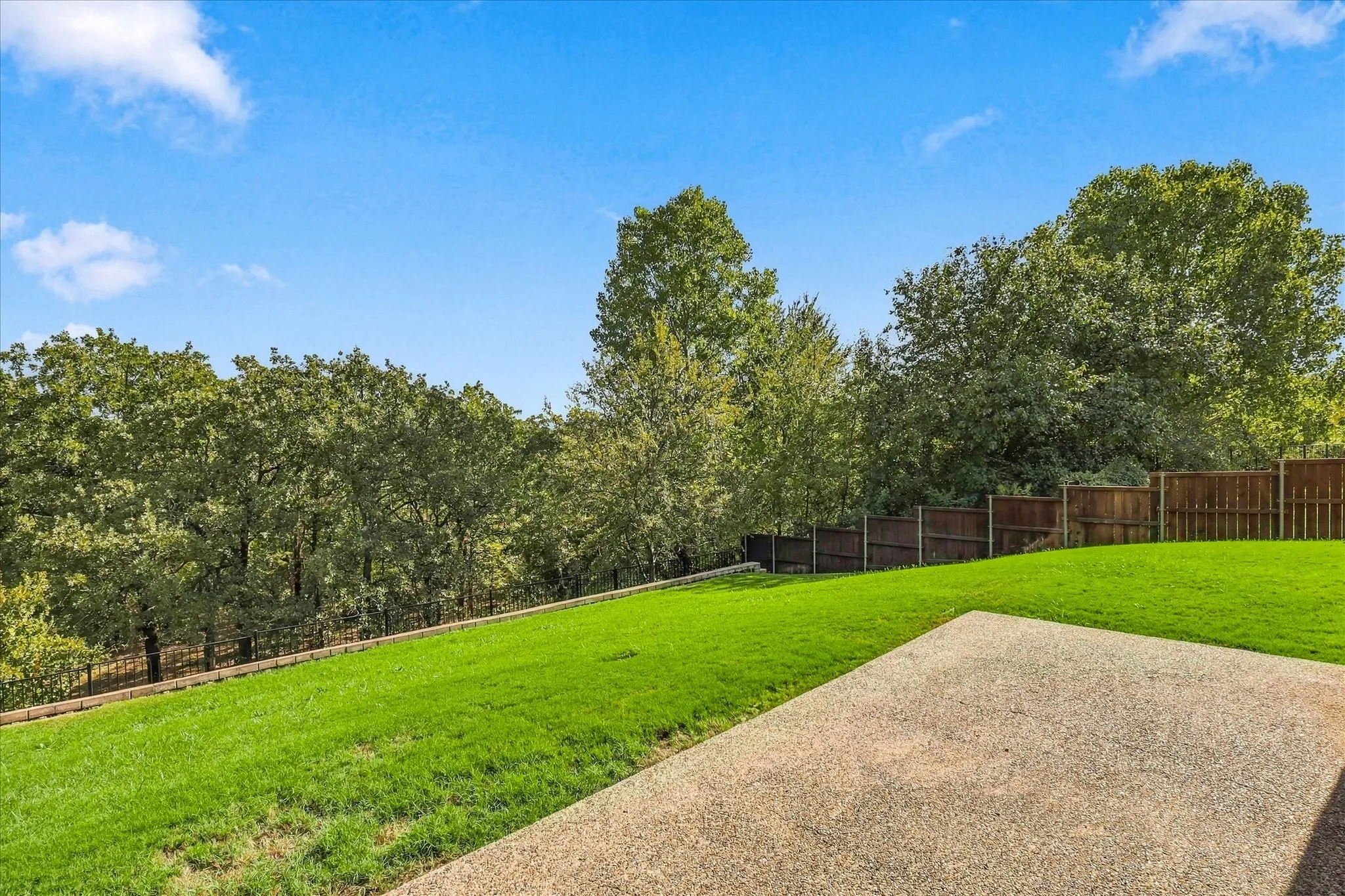Expansive backyard featuring a manicured lawn, exposed aggregate patio, wood privacy fencing, and mature trees
