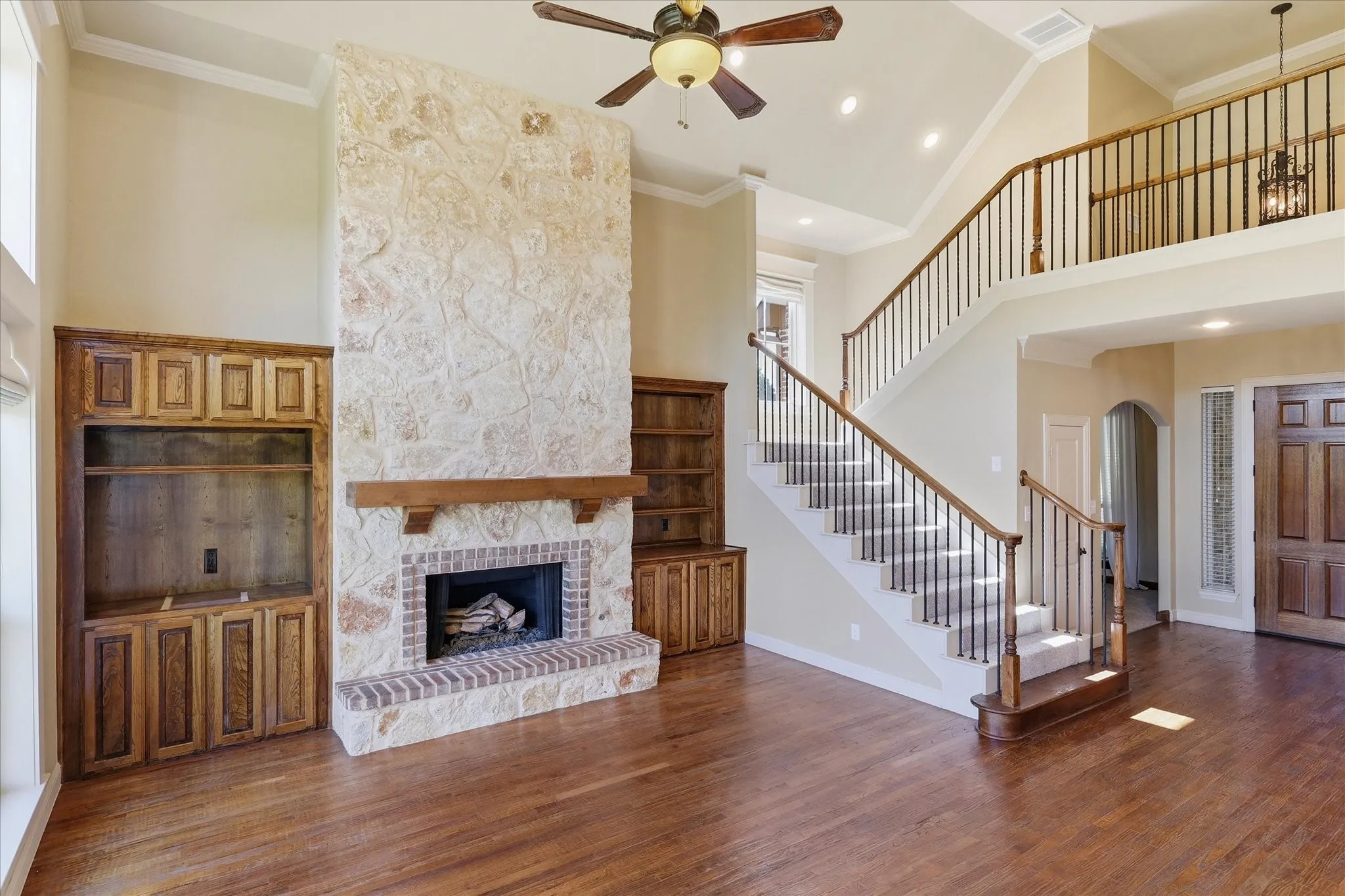 Floor-to-ceiling stone fireplace with wood mantel, custom built-in cabinetry, wood-finish flooring, vaulted ceiling, and staircase with wooden railing