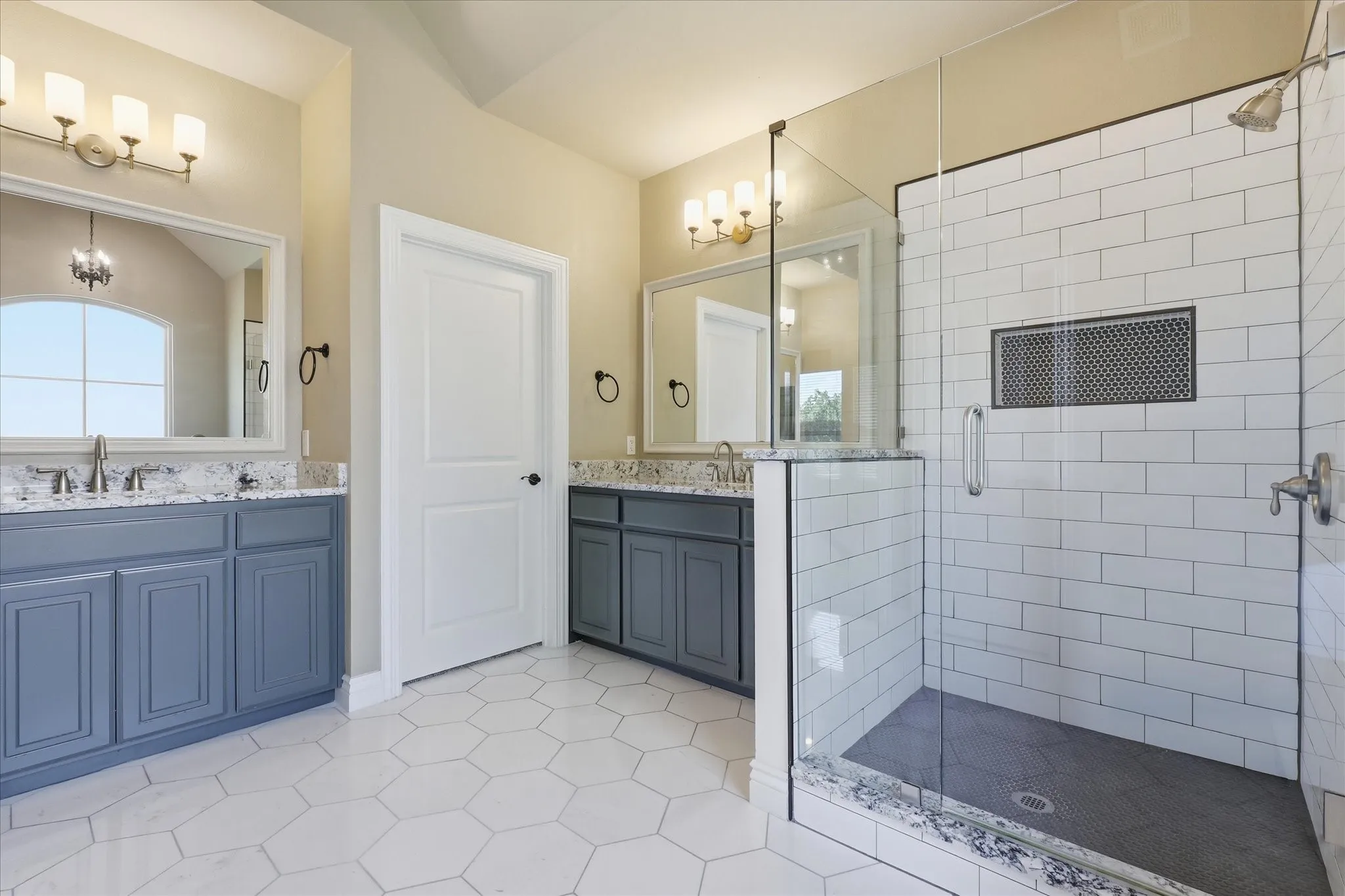 Dual blue-toned vanities featuring stone-finish countertops, glass-enclosed shower with subway tile surround, and hexagonal tile flooring