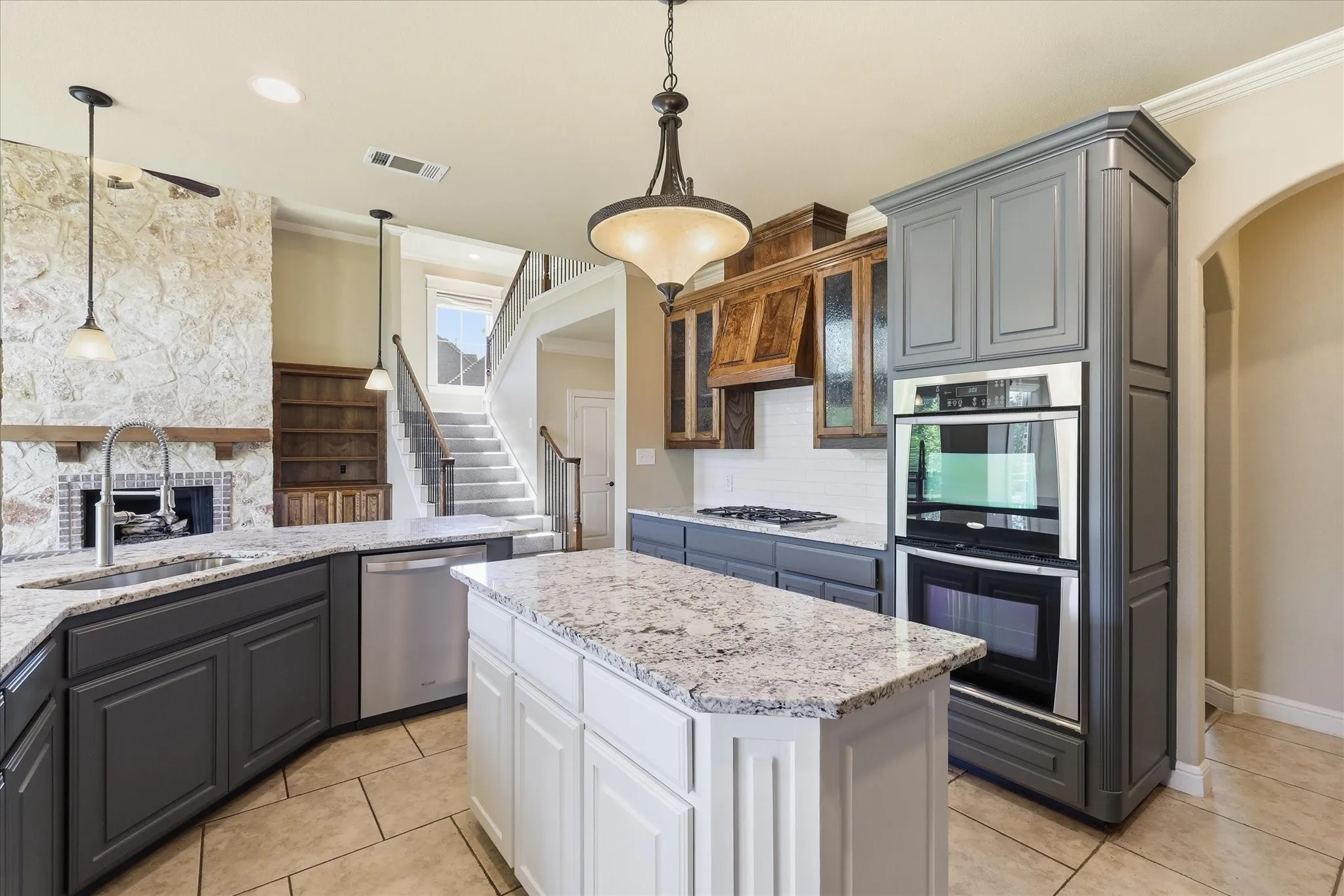 Kitchen featuring a stone-faced fireplace, center island with stone countertop, stainless steel double wall ovens, two-tone cabinetry, and wood-finish cabinetry accents