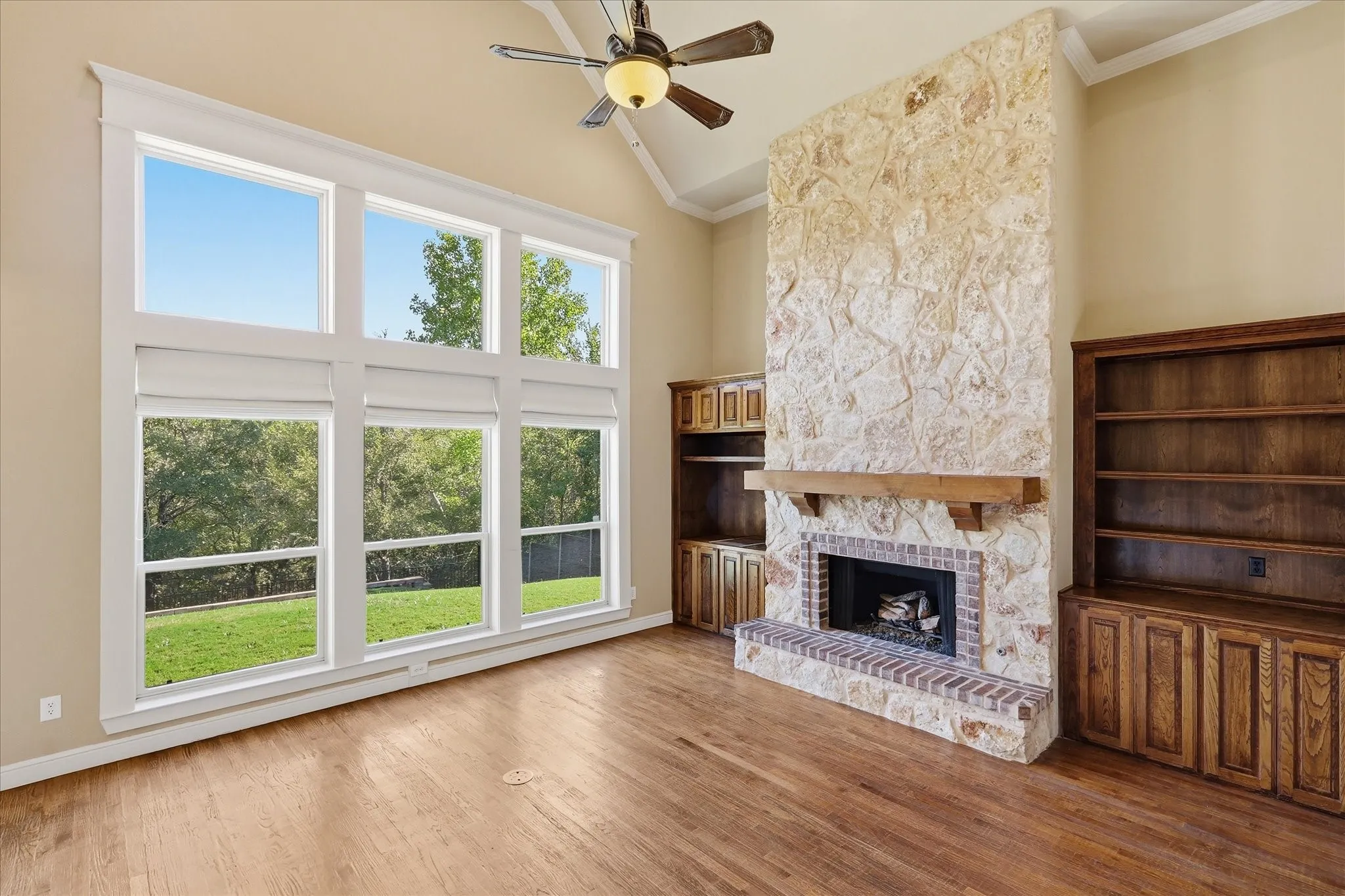 Vaulted ceiling with a central fan, large multi-pane windows, floor-to-ceiling stone fireplace with a wood mantel, and custom wood-finish built-in cabinetry
