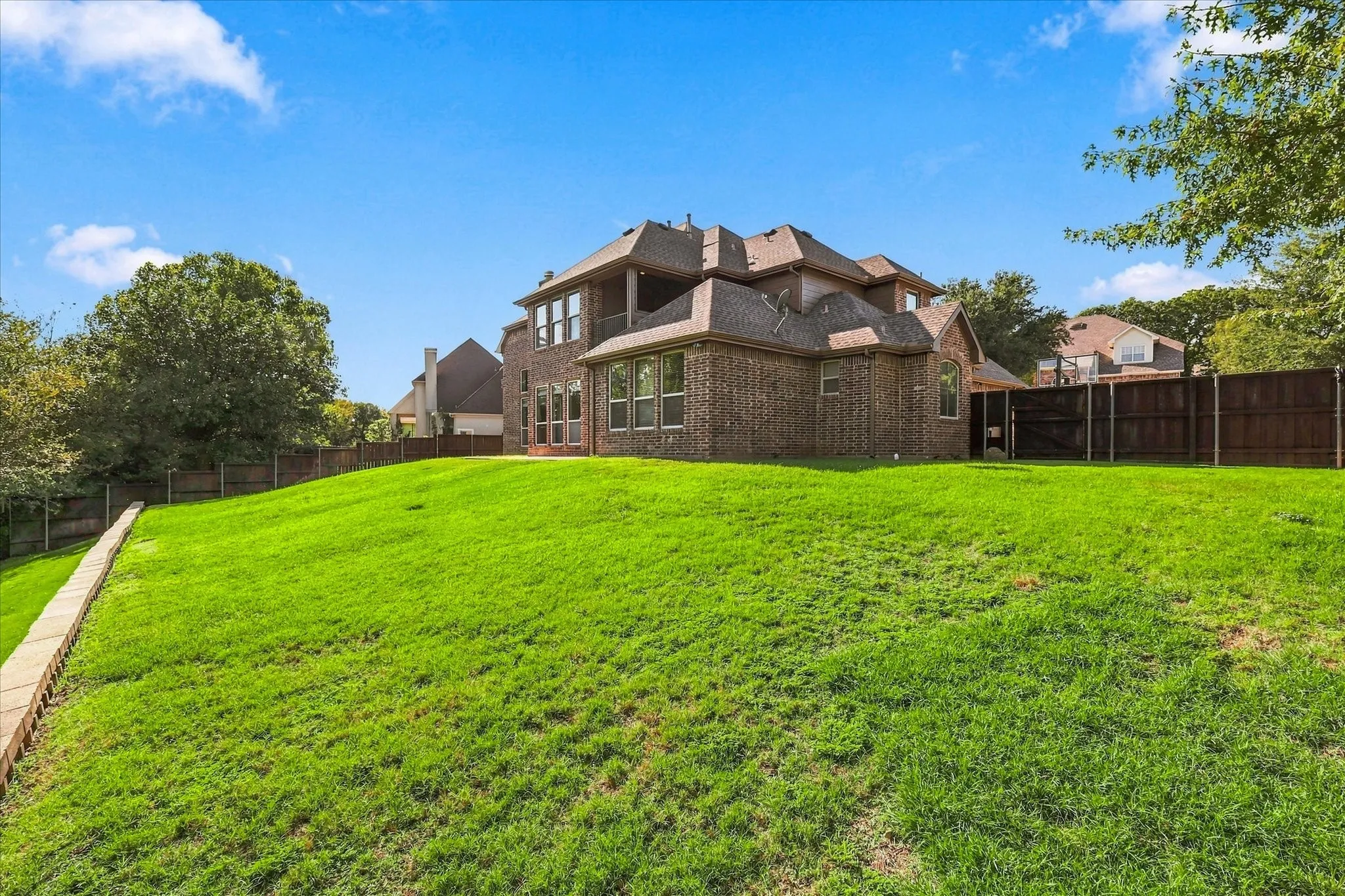 Brick exterior two-story residence featuring an expansive grass lawn, tiered stone retaining wall, and wood privacy fencing