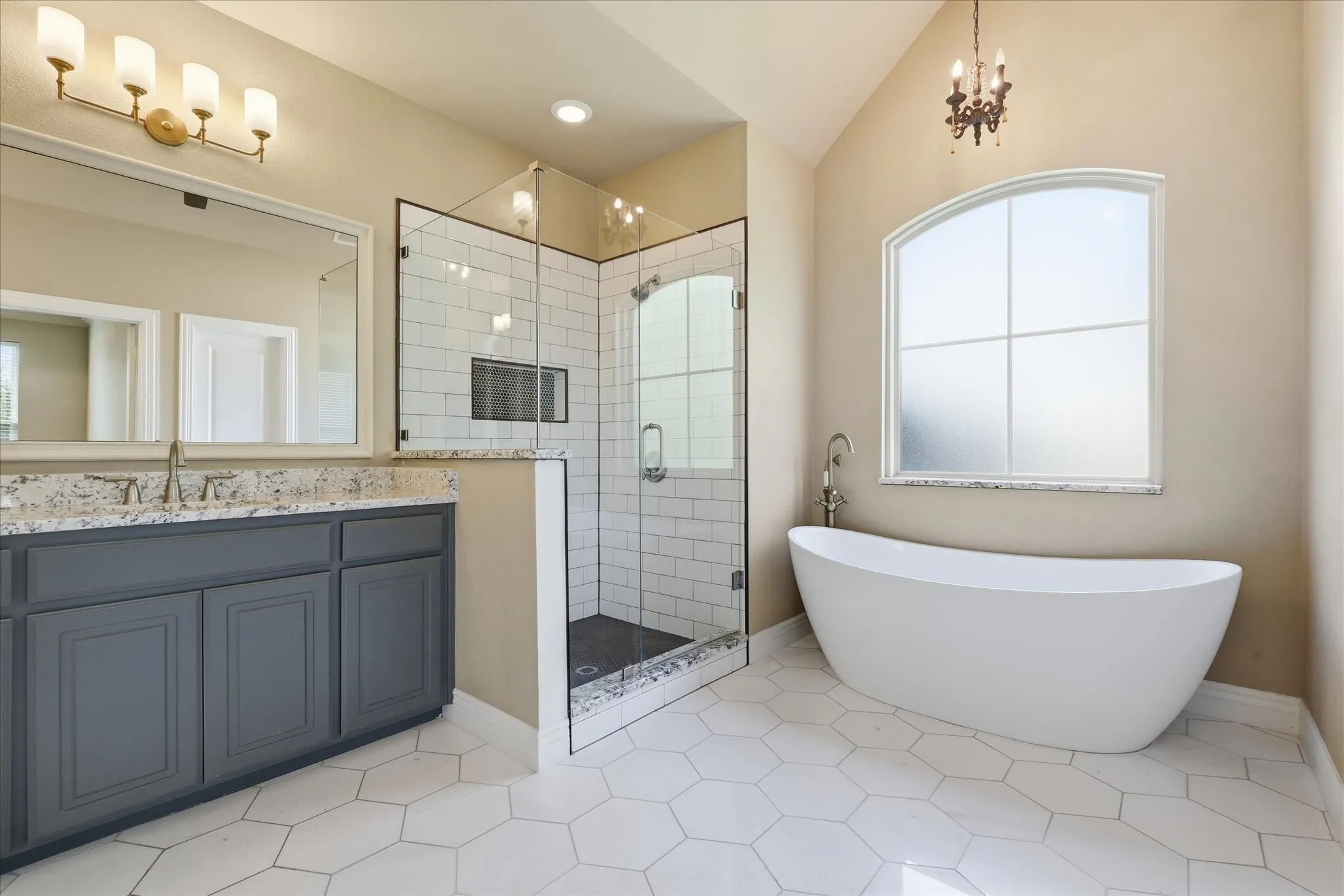 Freestanding soaking tub beneath an arched window, dual-sink vanity with stone countertop, and walk-in shower featuring a glass enclosure and subway tile surround, finished with hexagonal floor tile and vaulted ceilings