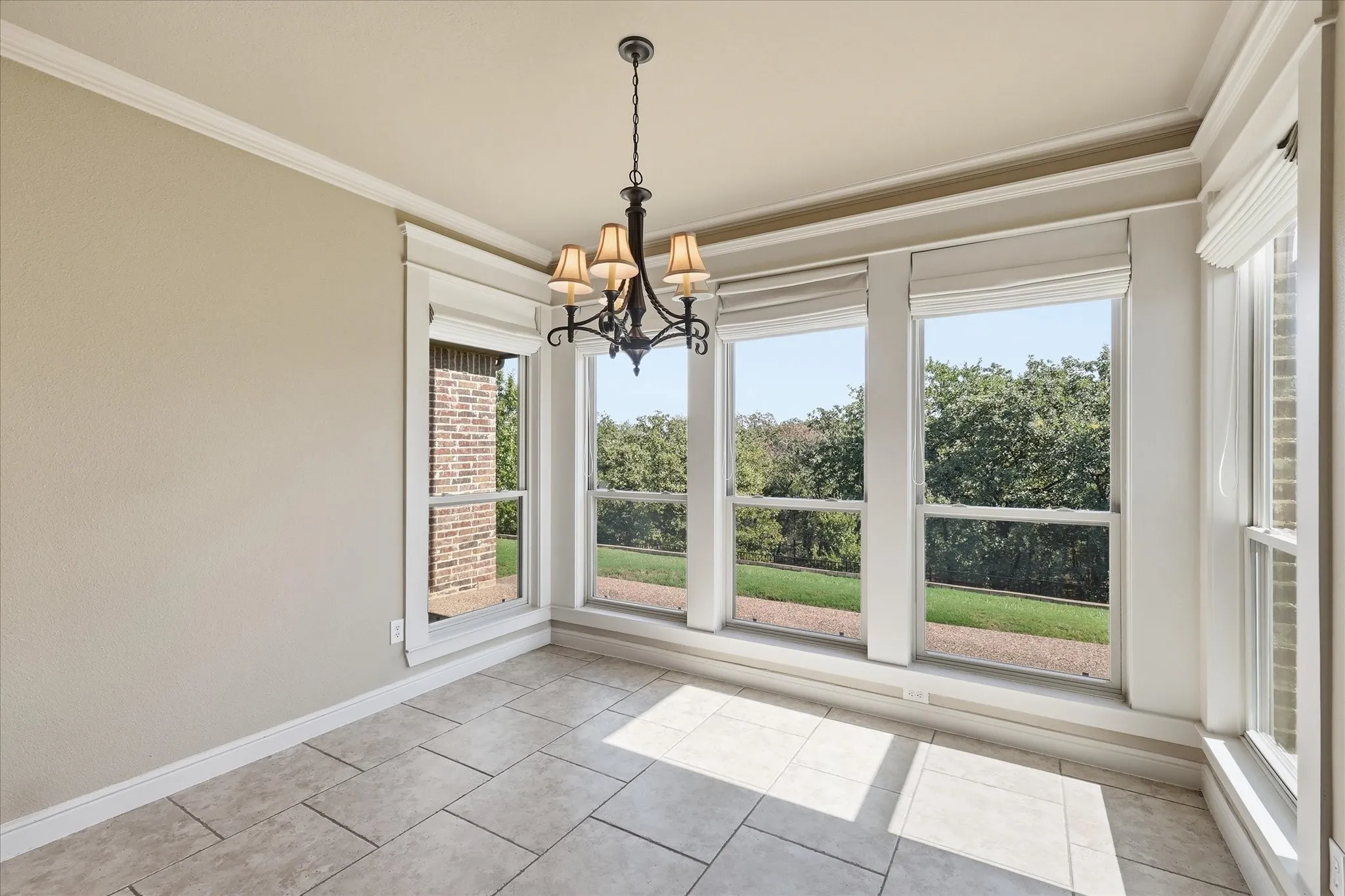 Natural light-filled room featuring large windows with window treatments, neutral stone-finish tile flooring, crown molding, and an intricate dark metal multi-arm chandelier